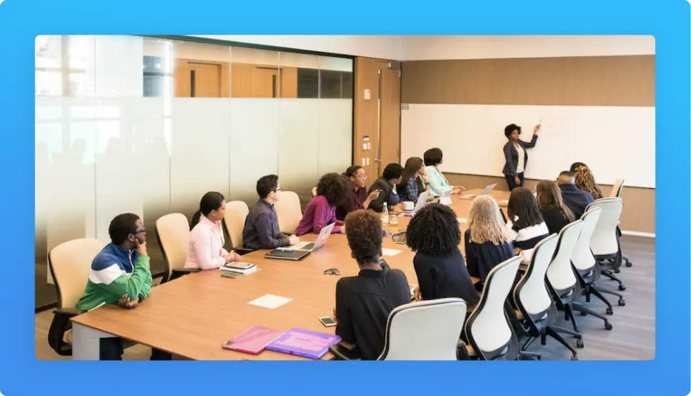Woman standing pointing a white board explaining a sale to a seated audience. Office mood