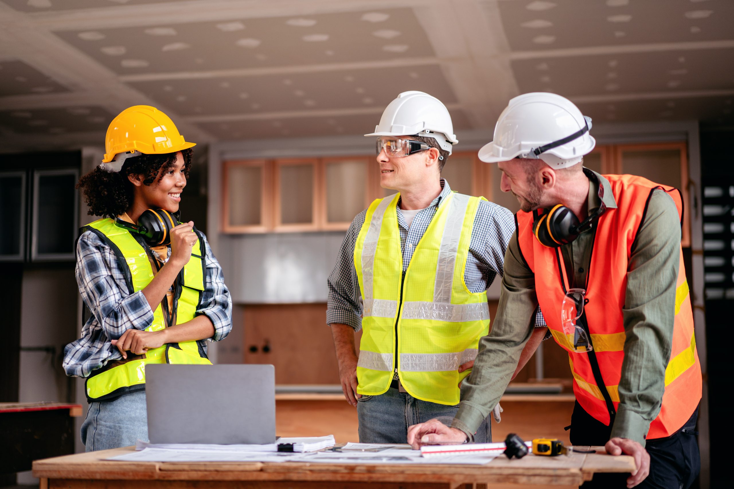 Three construction workers, two men and a woman, discuss plans at a worksite, wearing safety helmets and vests.