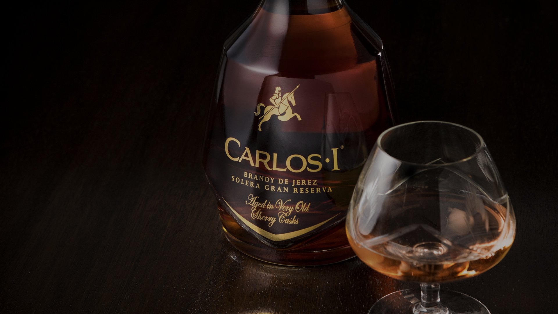 A dark amber brandy bottle labeled "Carlos I" stands beside a half-filled glass on a dark wooden surface, exuding elegance and sophistication.