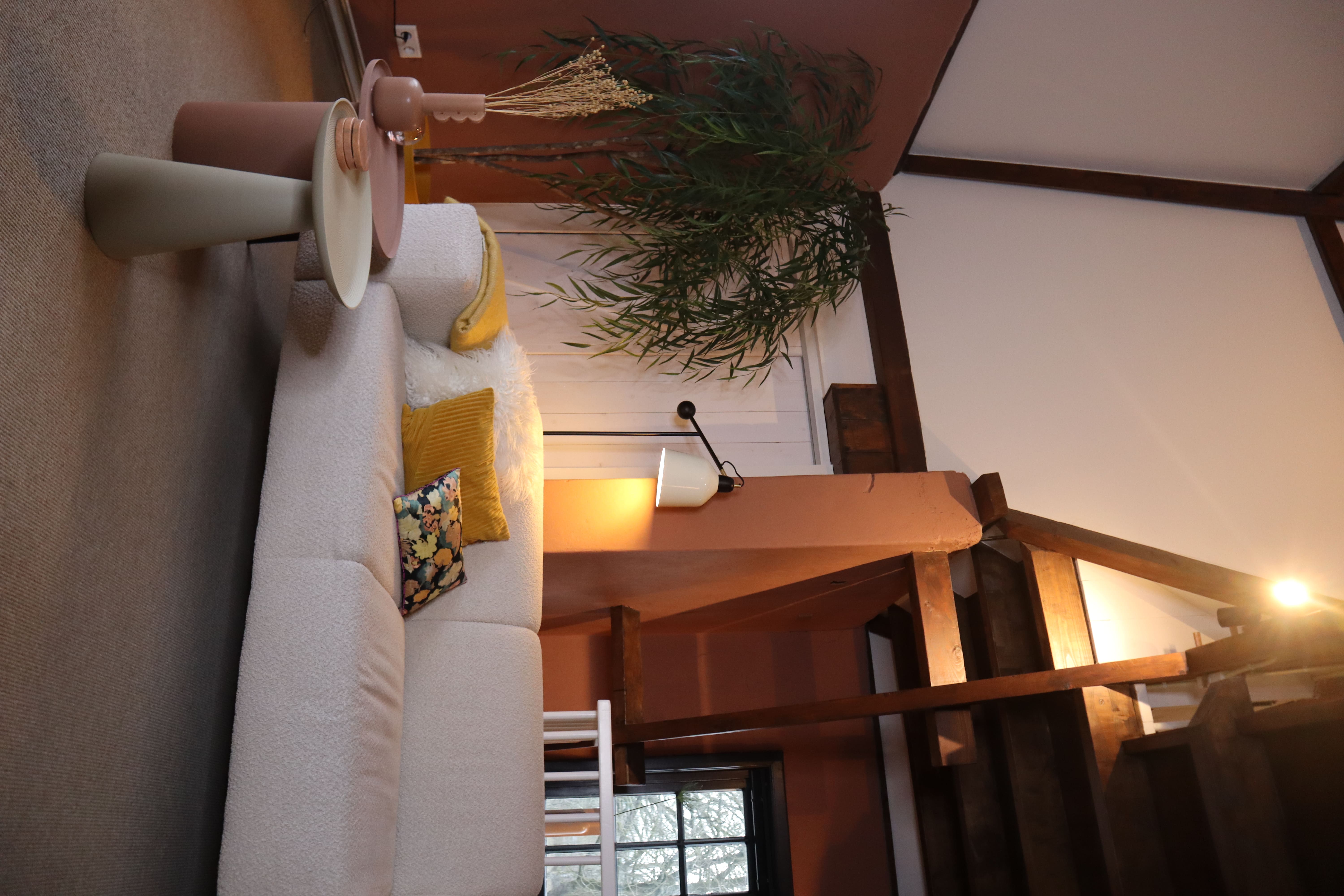 Gezellige woonkamer met een witte bank, pastelkleuren bijzettafels, een grote kamerplant in een gele pot, en zachte verlichting. De ruimte heeft een minimalistisch ontwerp met houten balken.