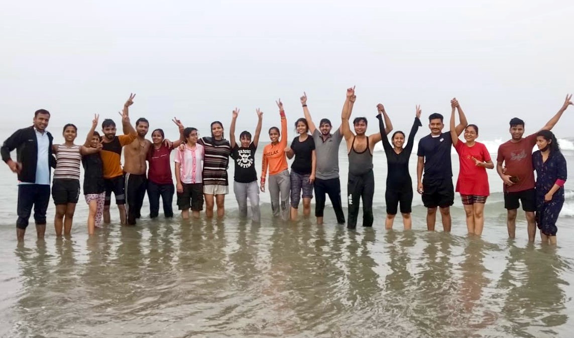 A smiling young man with crossed arms, wearing a plaid shirt and white t-shirt, poses against a dark background.