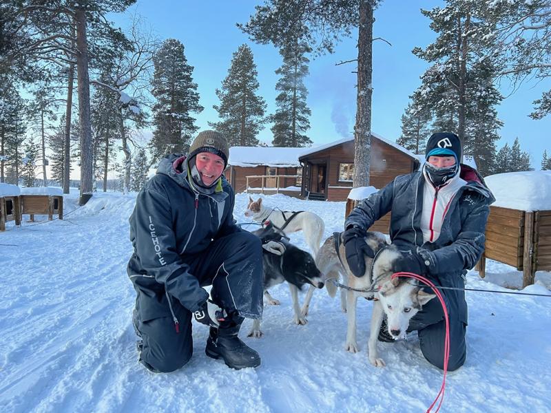 Two people are kneeling in the snow next to sled dogs in a snowy forest setting with a wooden cabin in the background.