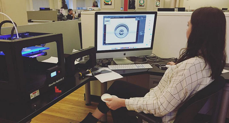 A photo of a woman working on a computer and checking a 3D schematic of an object. There is also a 3D printer near her workstation.