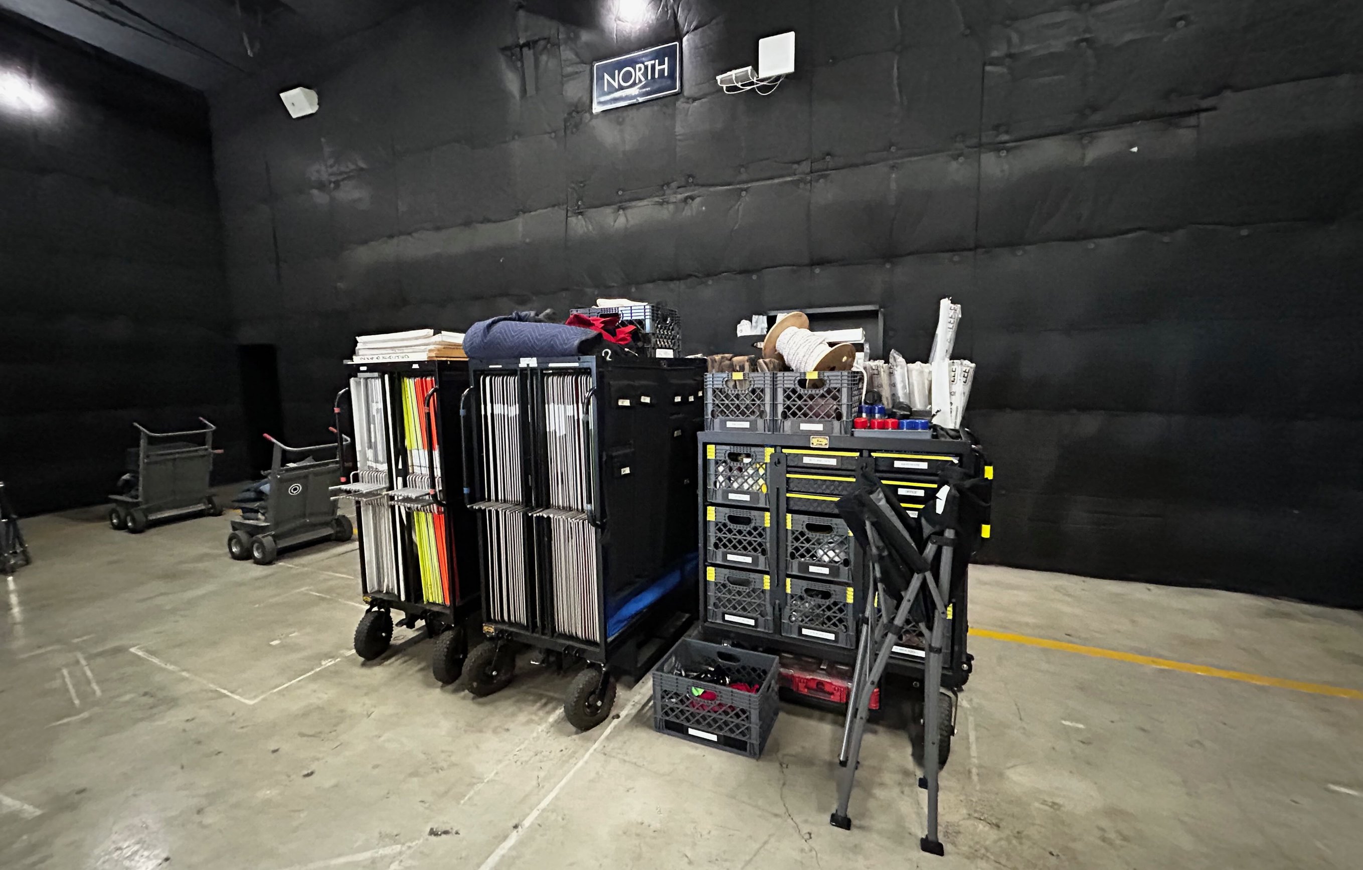 A grip package staged on a black-walled stage under a 'North' sign - a taco cart of vertical flags and frames, a frame cart, and a seven-crate set cart loaded with labeled milk crates, rope spools and apple boxes.