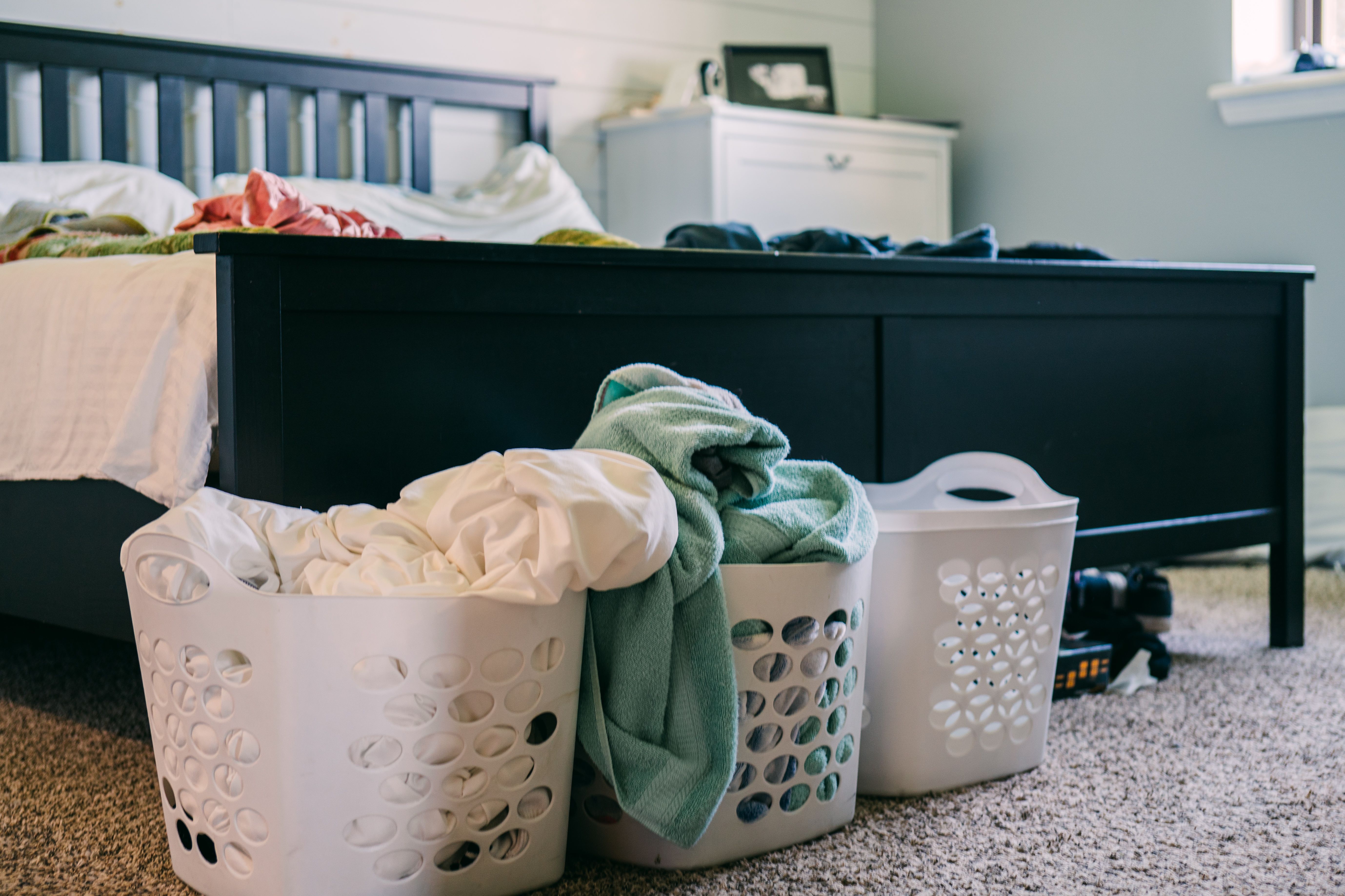 Three overflowing laundry baskets on the carpet beside an unmade, clothes-covered bed, the kind of messy bedroom that piles up with ADHD.