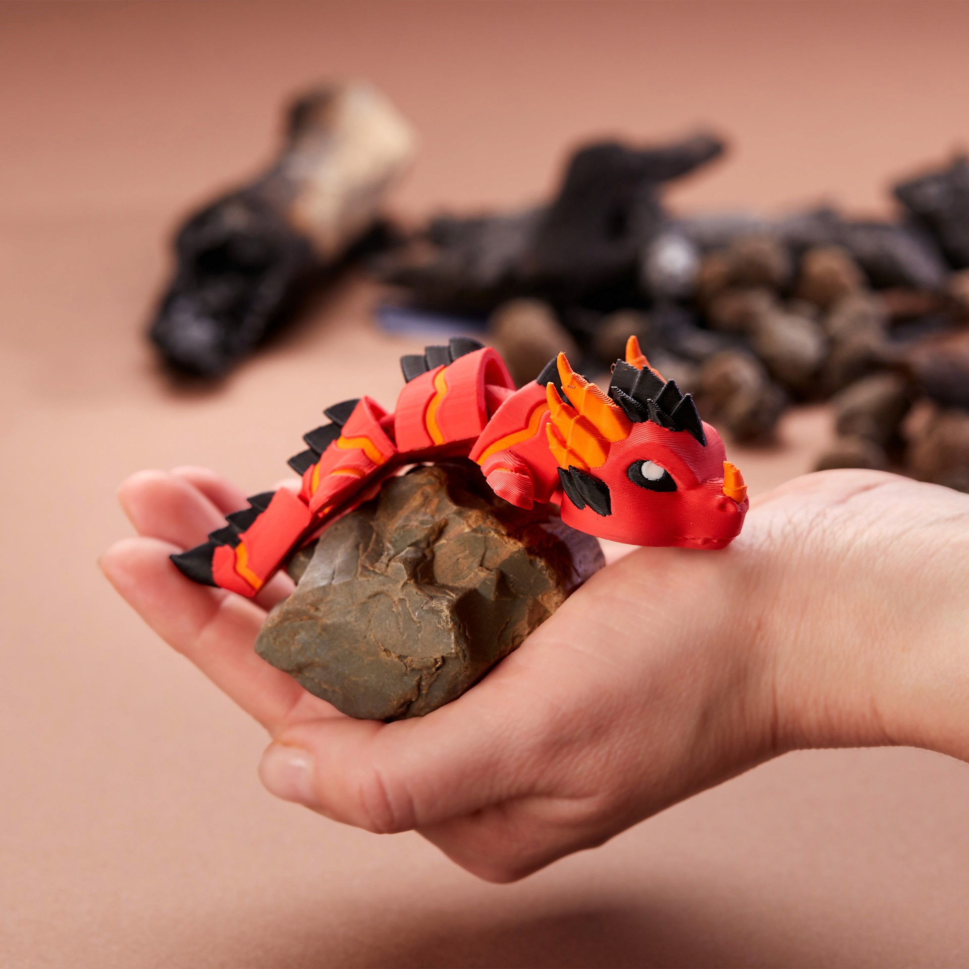A hand holds a detailed red and black 3D-printed dragon figurine with scale-like texture, set against a blurred background with stones.
