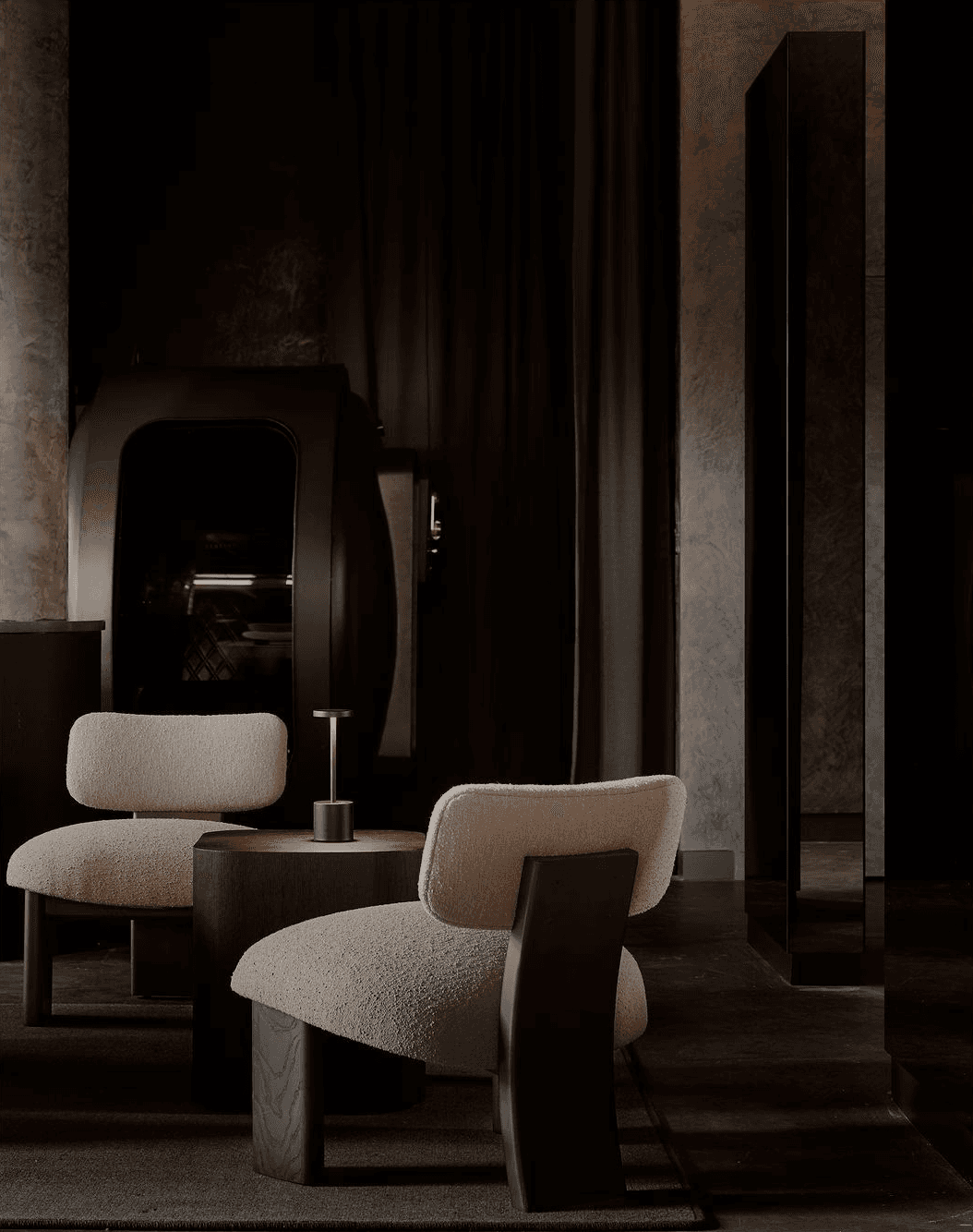 Dark lounge with two sculptural cream chairs, a small round table, and tall mirrors.