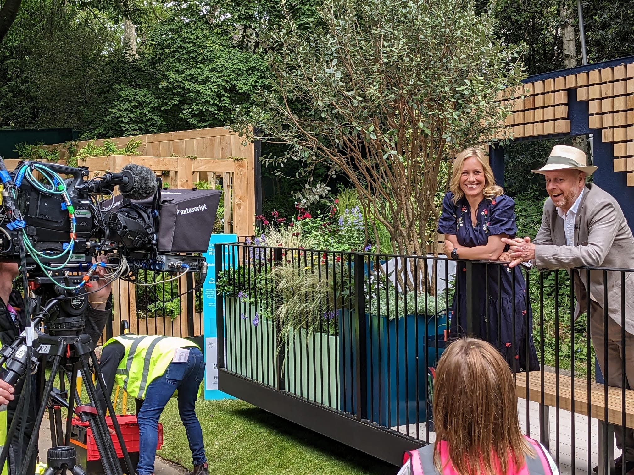 A person speaks outdoors near a garden display, while a film crew records the scene with cameras and equipment.