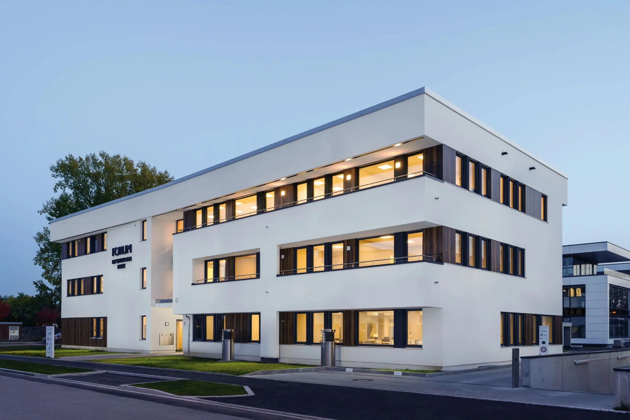 Modernes Bürogebäude mit weißen Fassaden, dunklen Fensterrahmen und beleuchteten Innenräumen, umgeben von gepflegten Grünflächen.