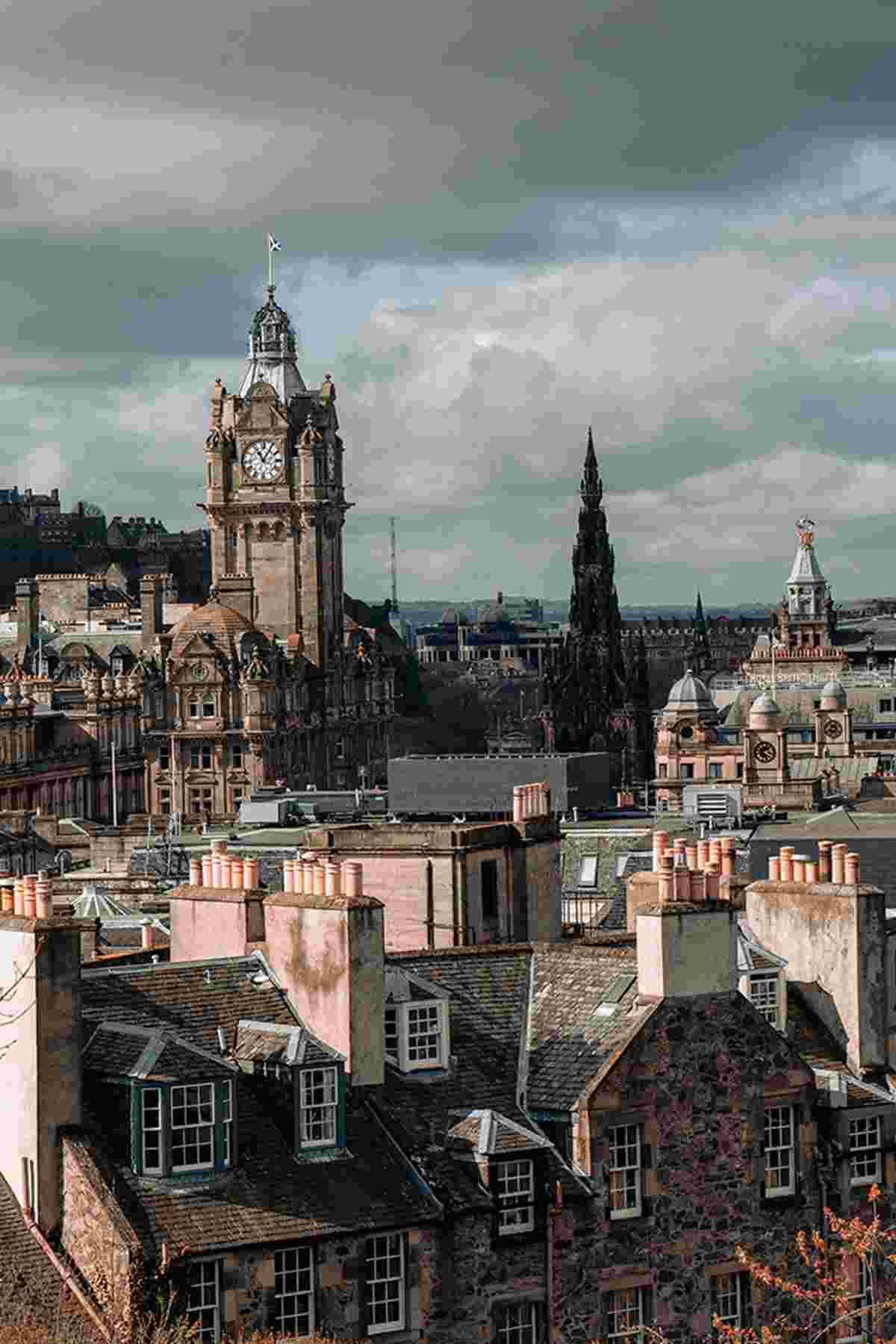 A cityscape featuring historic stone buildings and rooftops with chimneys in the foreground. Prominent landmarks, including a tall clock tower and gothic spire, rise against a cloudy sky.