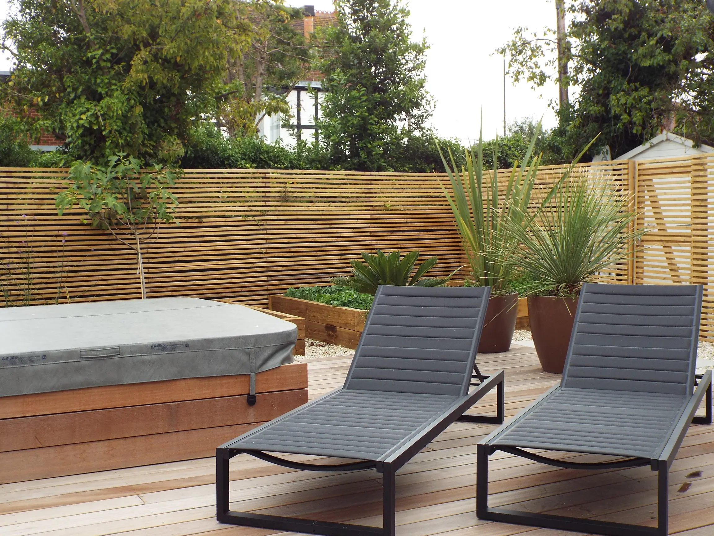 Two modern lounge chairs on a patio surrounded by greenery and wooden fencing, inviting relaxation.