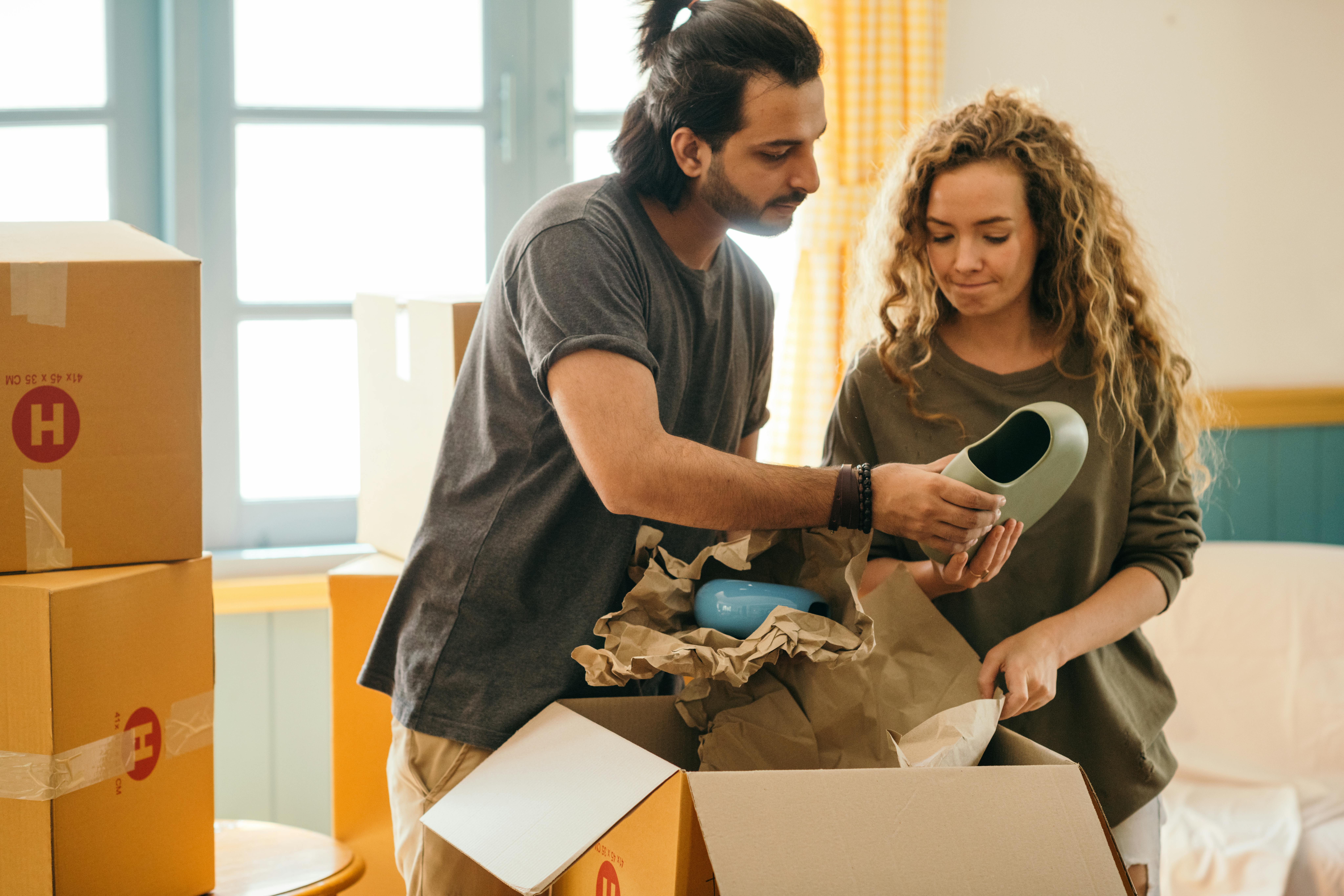 A newcomer couple in a bright 1-bedroom apartment unpacking cardboard delivery boxes and home essentials during their first week in Canada.
