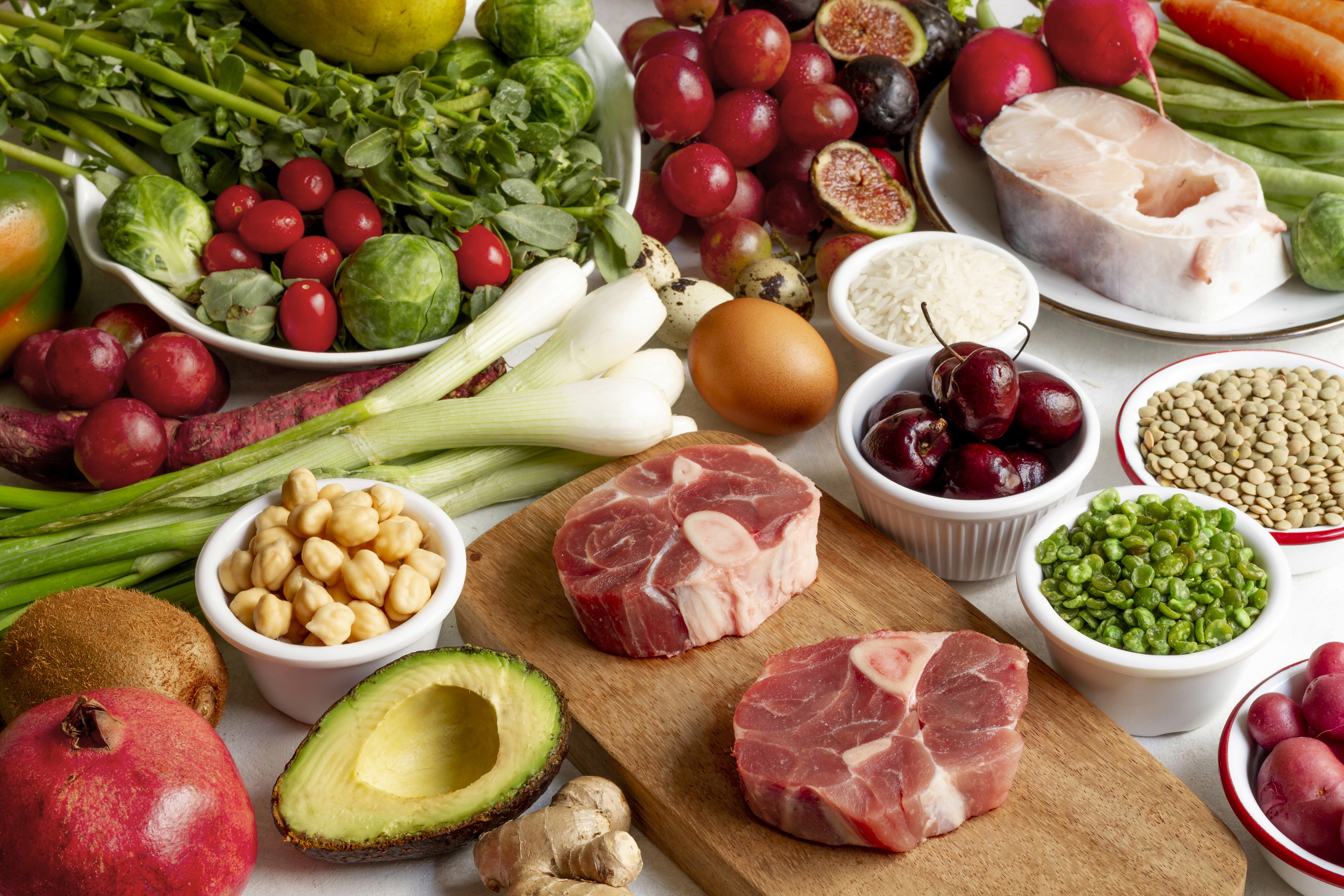 A colorful array of fresh foods on a table includes raw meat, fruits, vegetables, lentils, and grains. The setting is vibrant, suggesting health and abundance.
