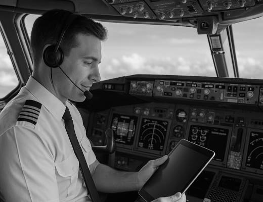 Commercial airline pilot wearing a headset, sitting in the cockpit and holding a tablet device while looking at the aircraft’s controls.