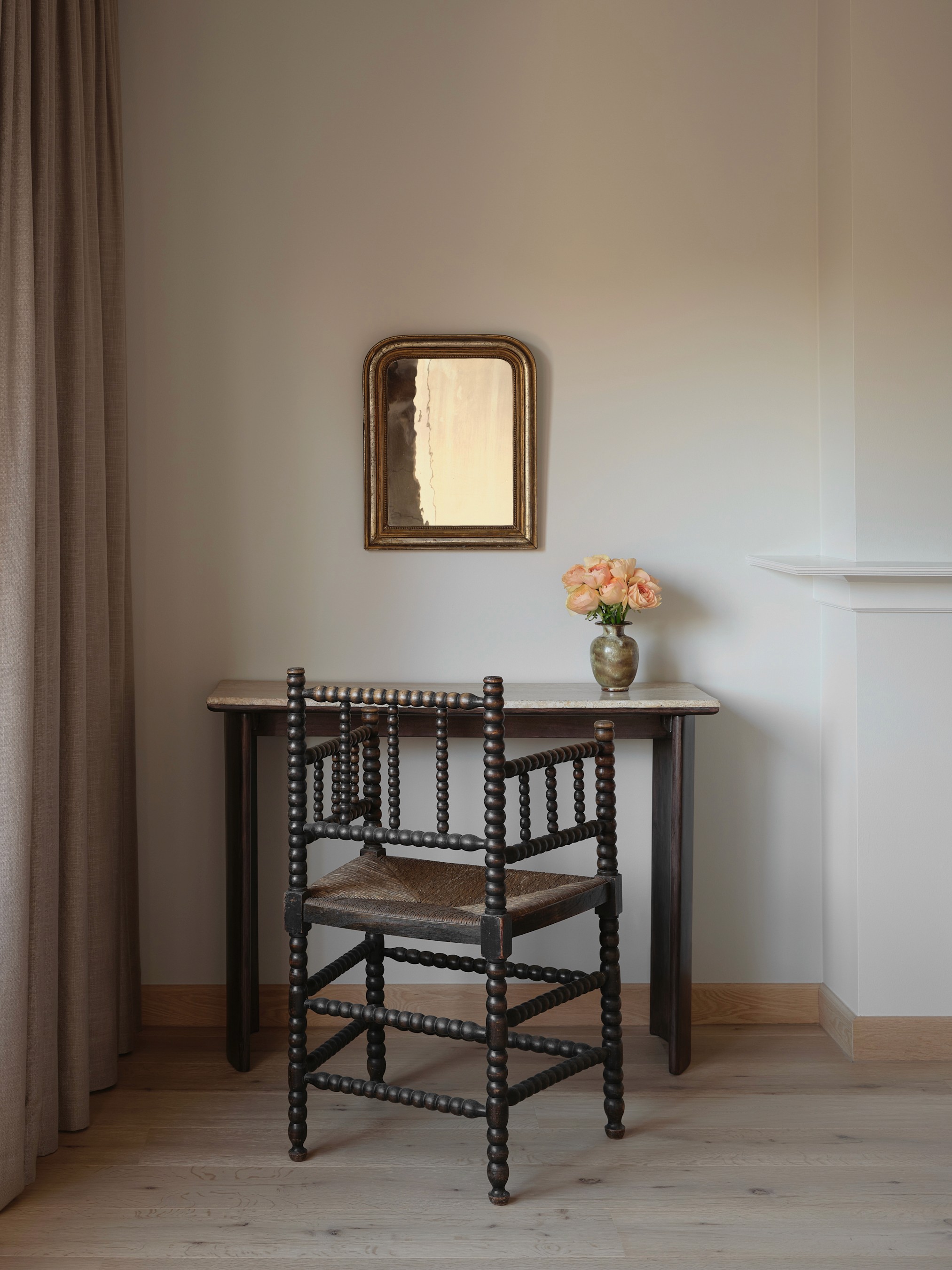 Bedroom corner with antique spindle chair, gilt-framed mirror, and dark wood and travertin console table with fresh roses