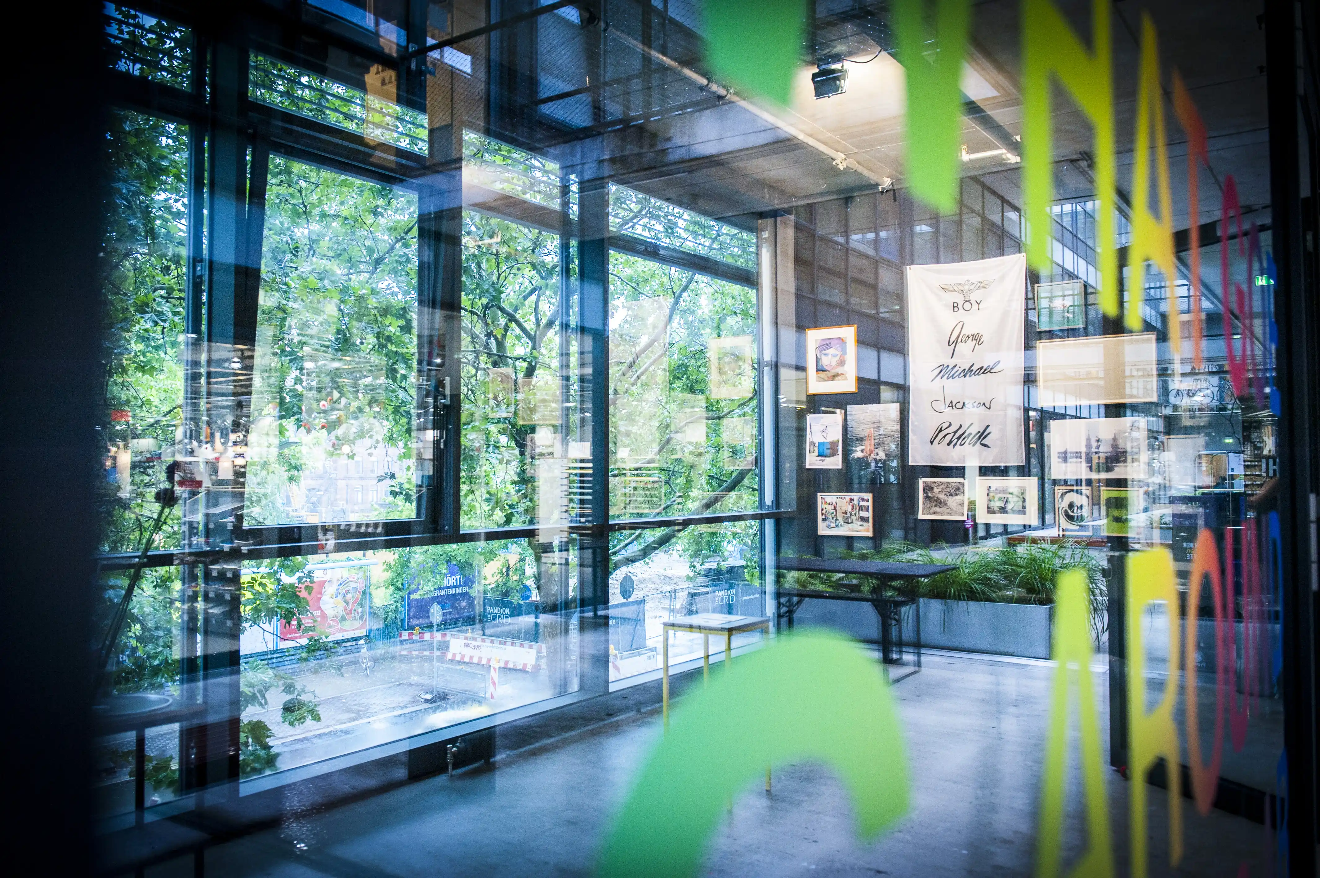 View through the glass facade of the STUDIOLO gallery with reflections of trees, artworks on the walls and a banner with artist signatures.