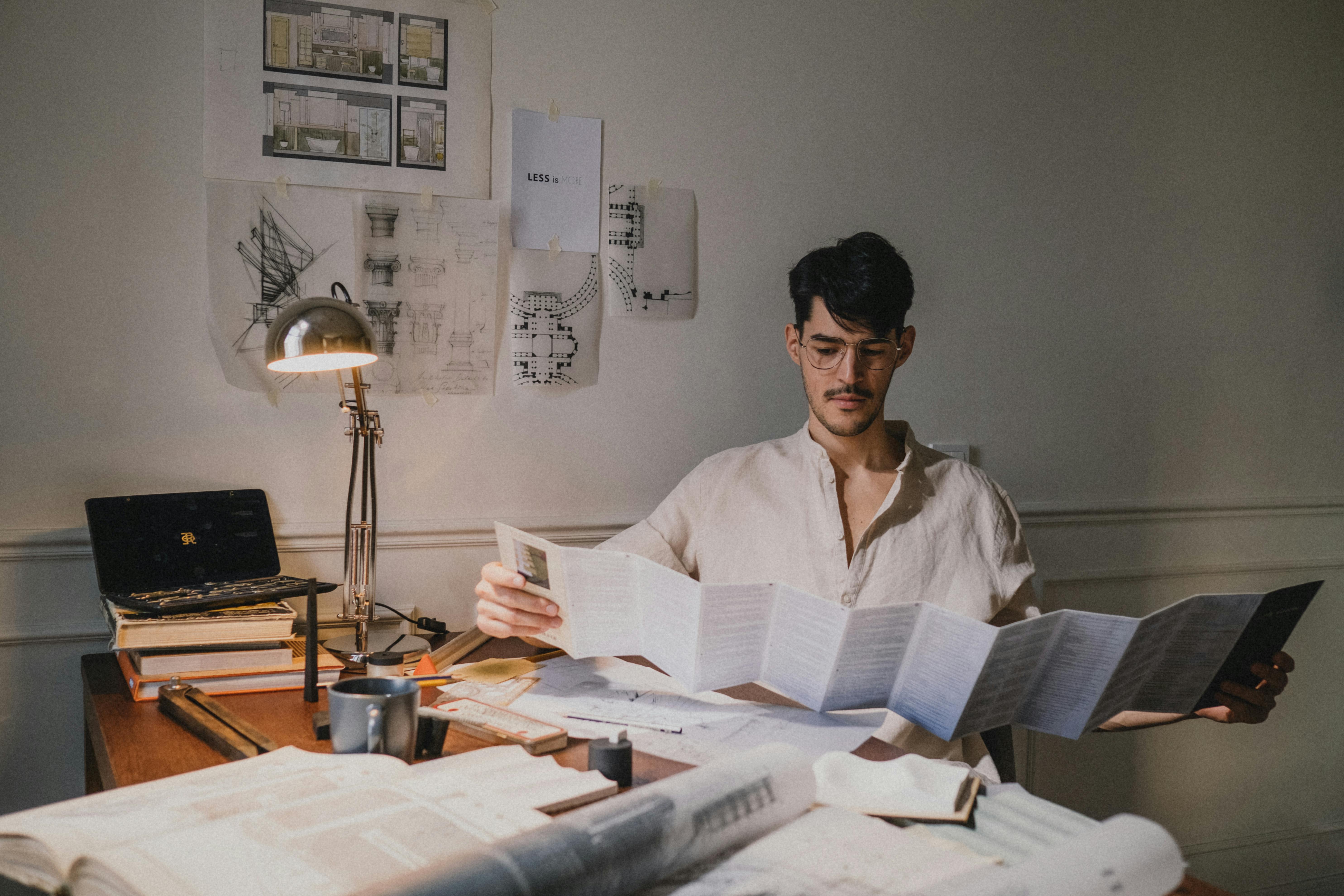 Man with glasses reviewing blueprints/papers at a cluttered desk.