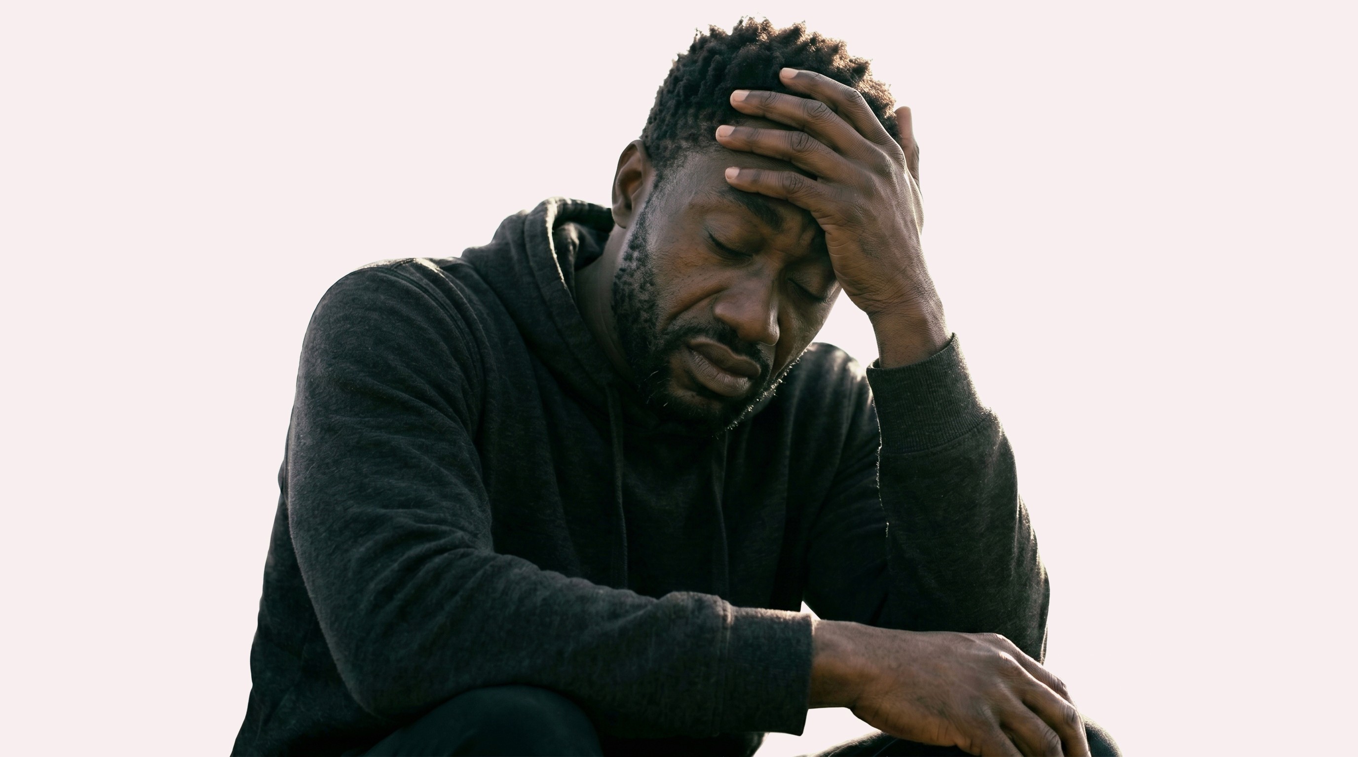 A person in a dark hoodie sits outdoors with a distressed expression, hand on forehead, conveying a sense of contemplation or stress against a plain background.