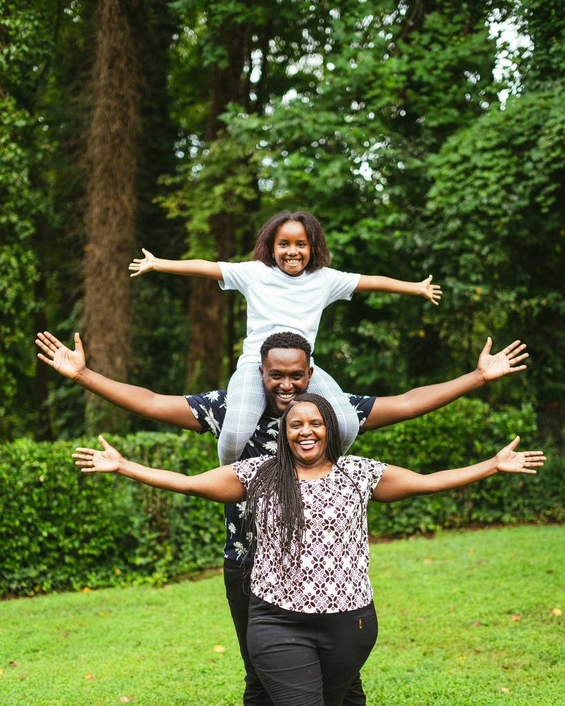 A joyful family posing playfully in a park, with the mother in front, the father behind her carrying their daughter on his shoulders, all spreading their arms wide and smiling amidst lush green surroundings.