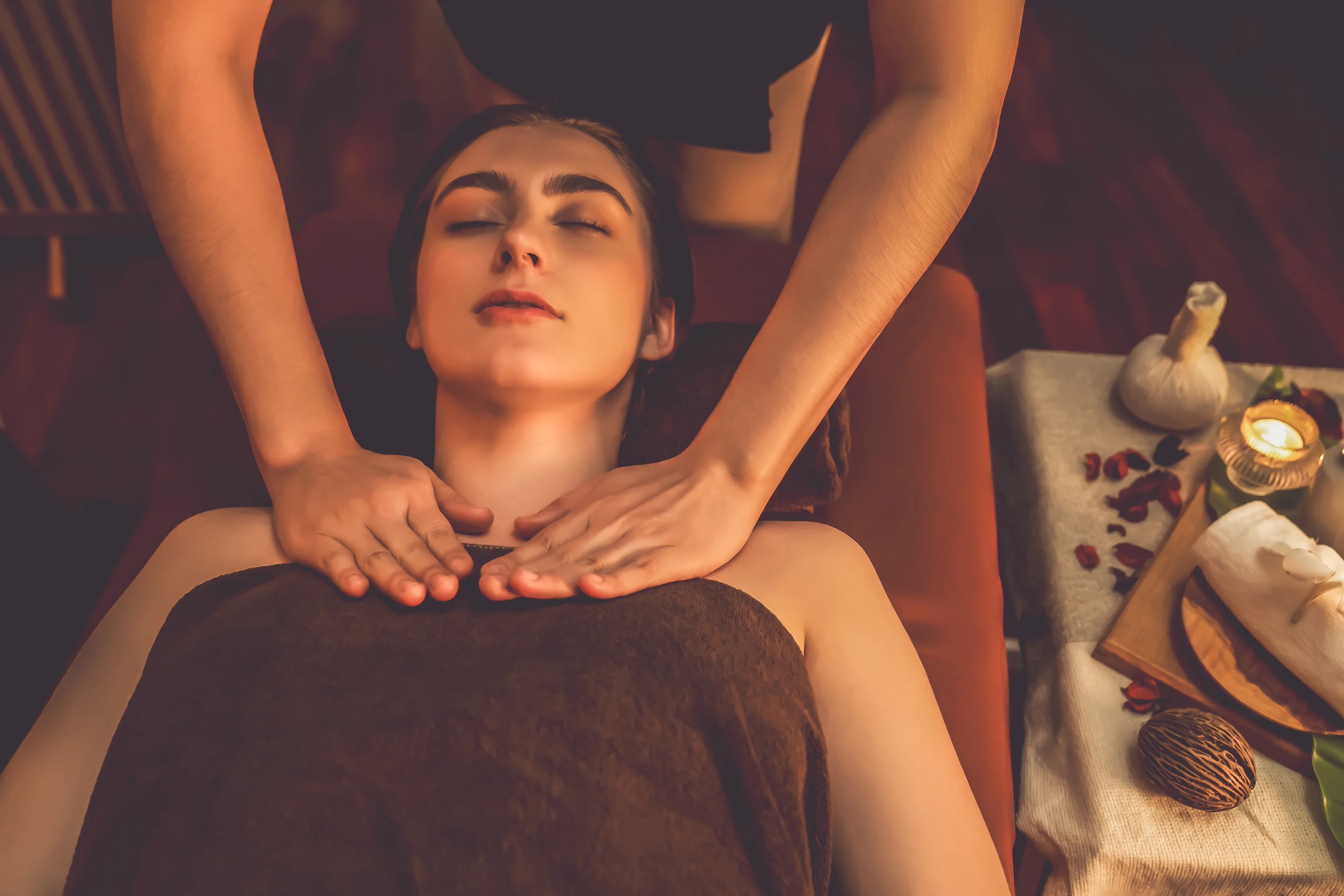 A woman receiving a relaxing Ayurvedic neck and shoulder massage in a warm, serene therapy room at Au Revoir Resort, Palakkad, Kerala, with herbal compresses, candles and wellness elements arranged beside her.