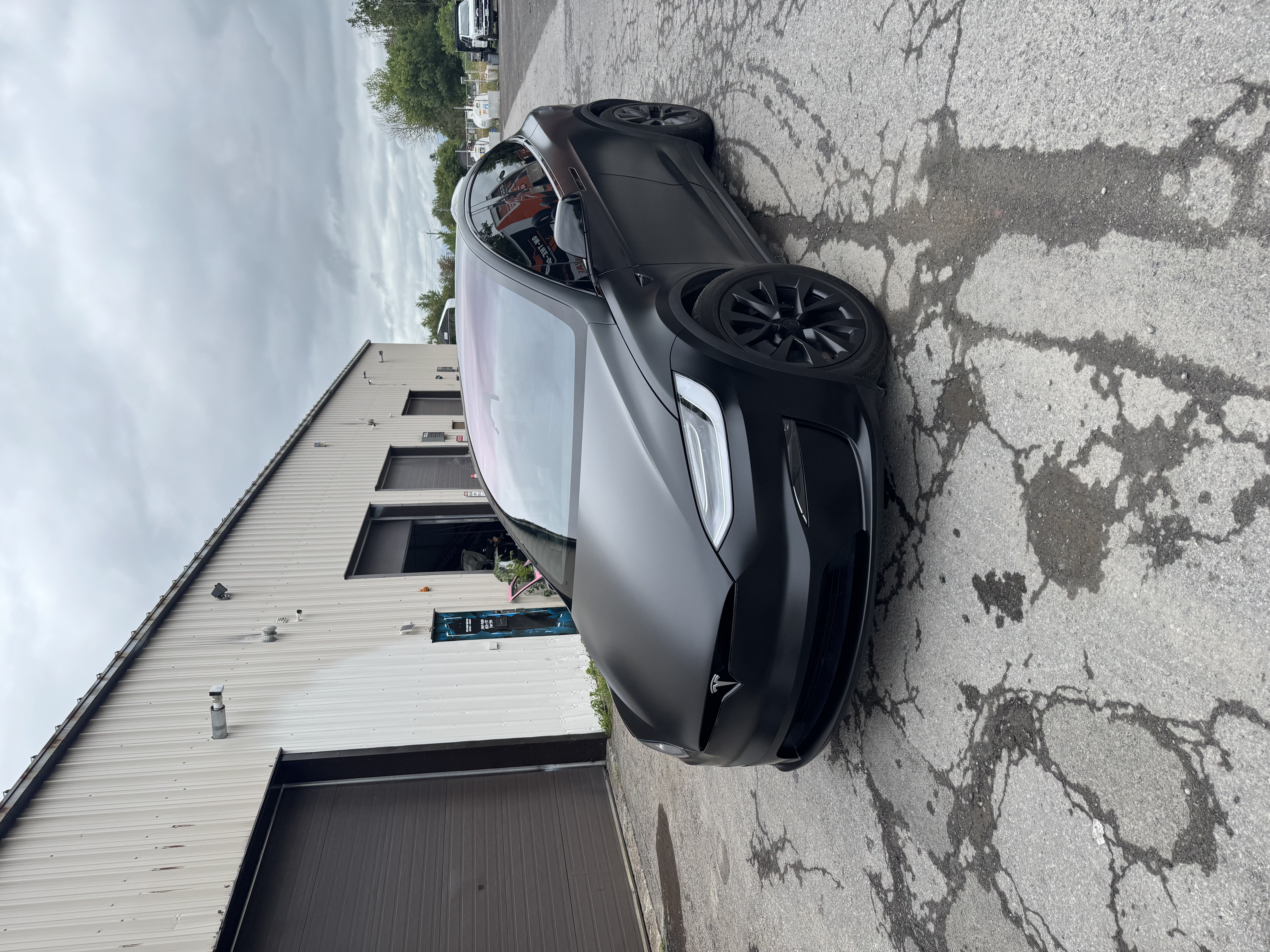 Matte black wrapped Tesla Model X with black wheels parked outside an automotive wrap shop.