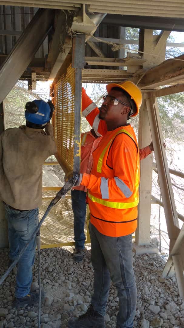 A man in a brown blazer and a woman in a light sweater sit at a desk with a laptop, discussing something. The room is dimly lit, creating a focused atmosphere.