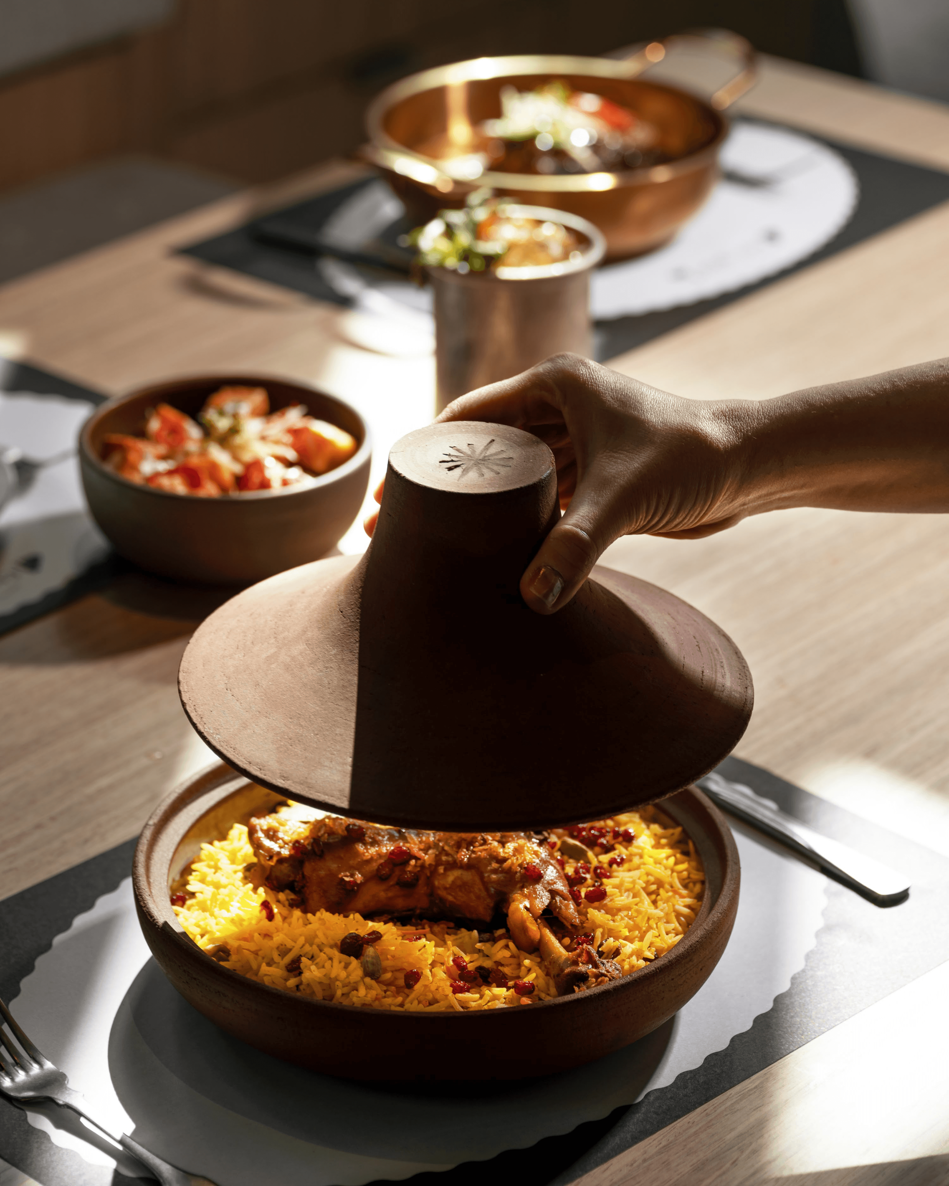 Hand lifting terracotta tagine lid from yellow saffron rice with chicken, red peppers beside copper bowls of vegetables and cocktail on East by West restaurant table.