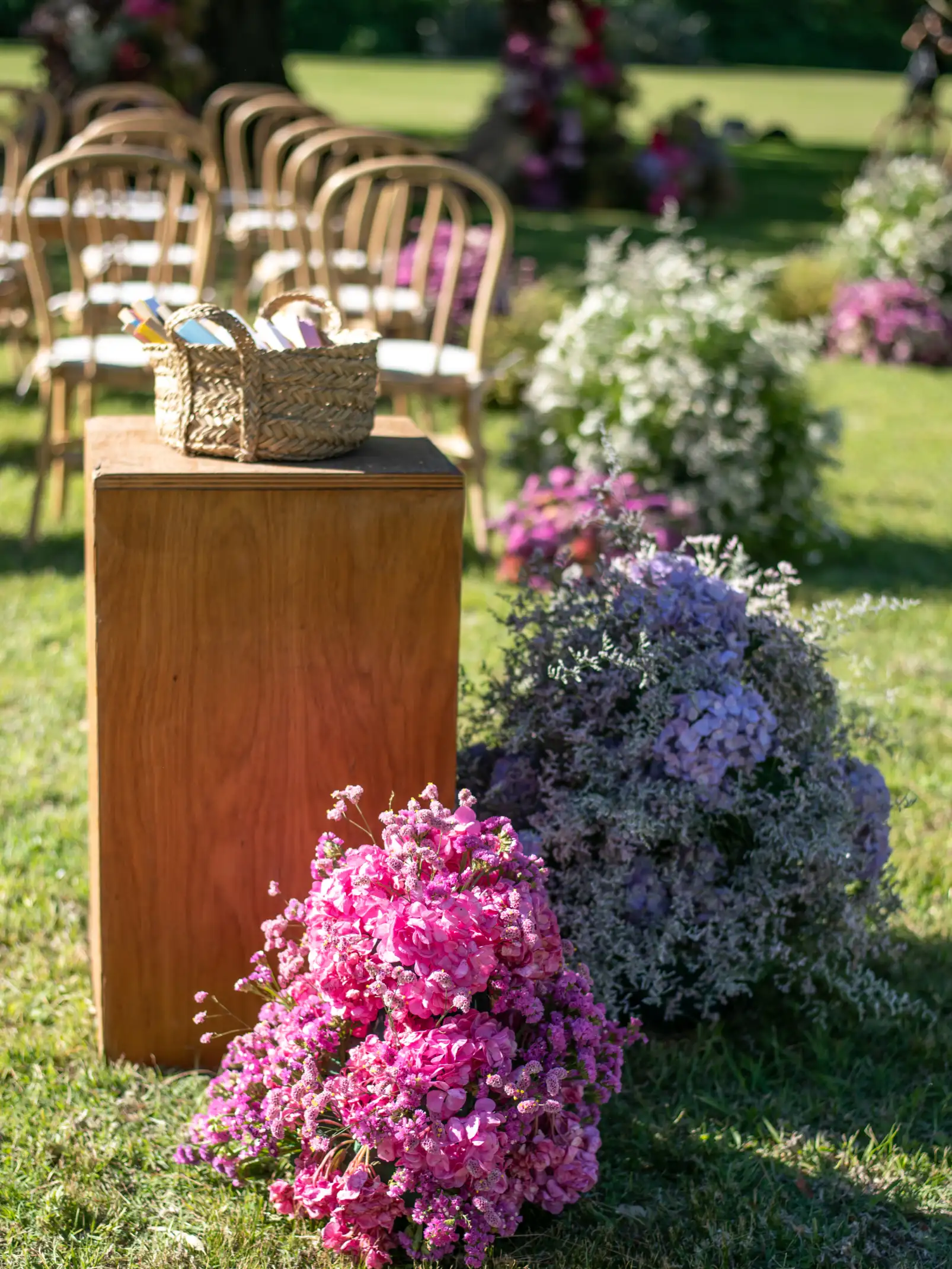 Entrada a ceremonia con flores en tonos rosas y lilas junto a pedestal de madera con canasta tejida.