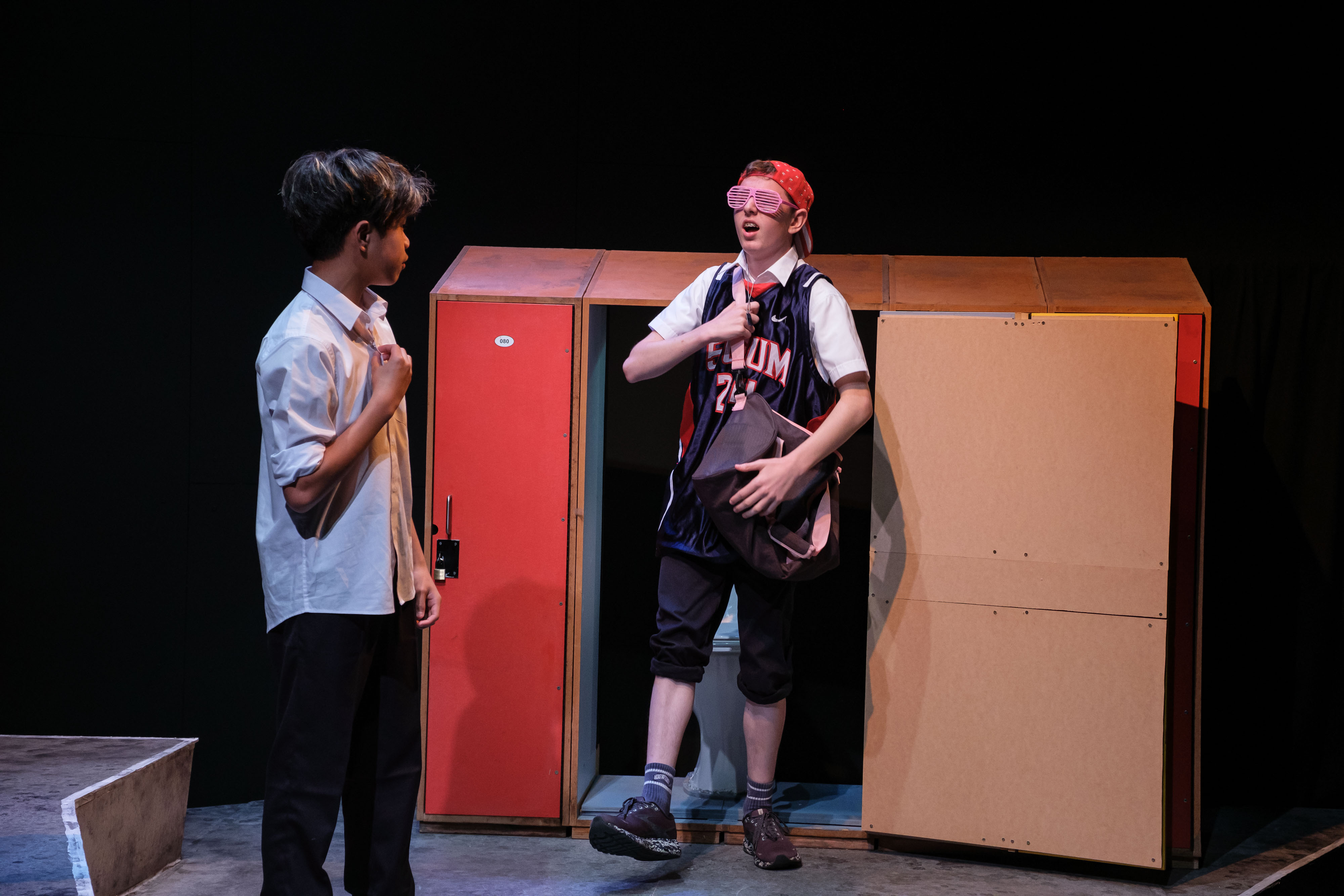 A student steps out of a locker wearing a basketball jersey and novelty shutter shades, surprising another student in a school uniform watching him. The exaggerated entrance appears to be a comedic moment within the school setting.