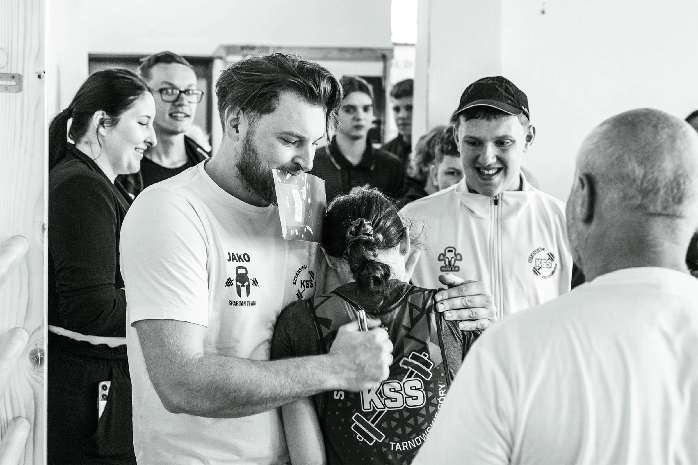 Czarno-białe, pełne emocji zdjęcie grupy osób w koszulkach Sztangiści KSS, przedstawiające wspólne świętowanie lub gratulacje