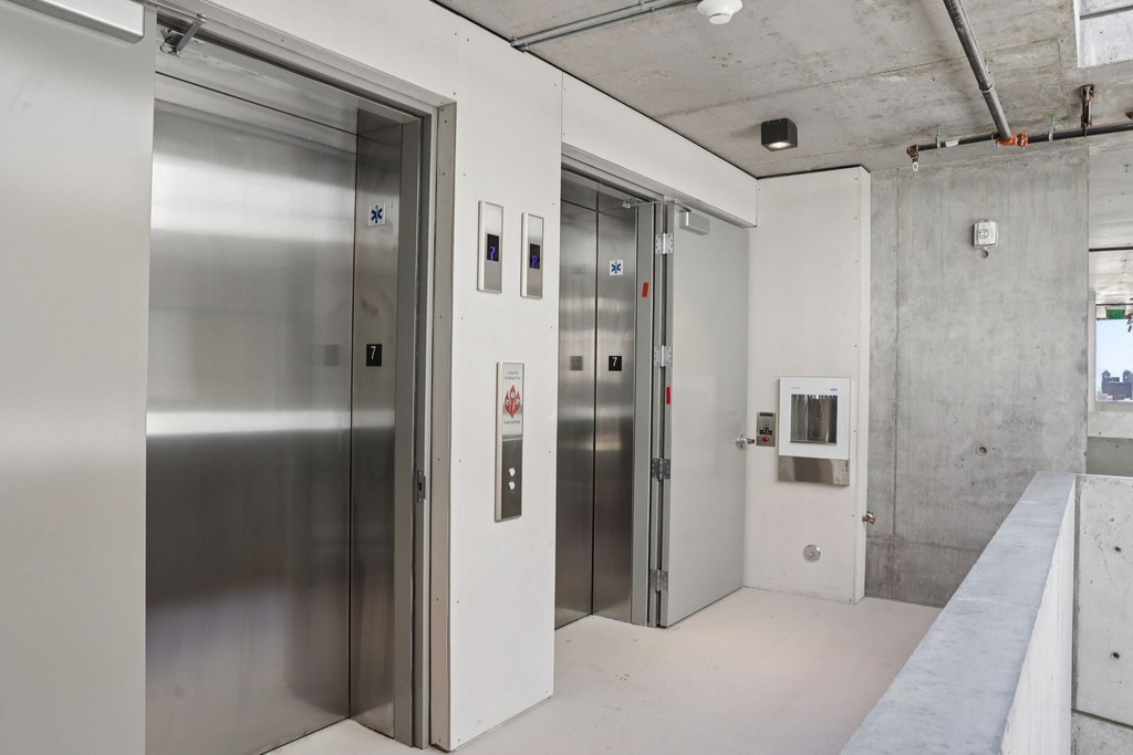 Two stainless steel elevators labeled 7 and 1 in a modern, concrete-walled building lobby. Bright, minimalistic, with overhead lighting and a small window.