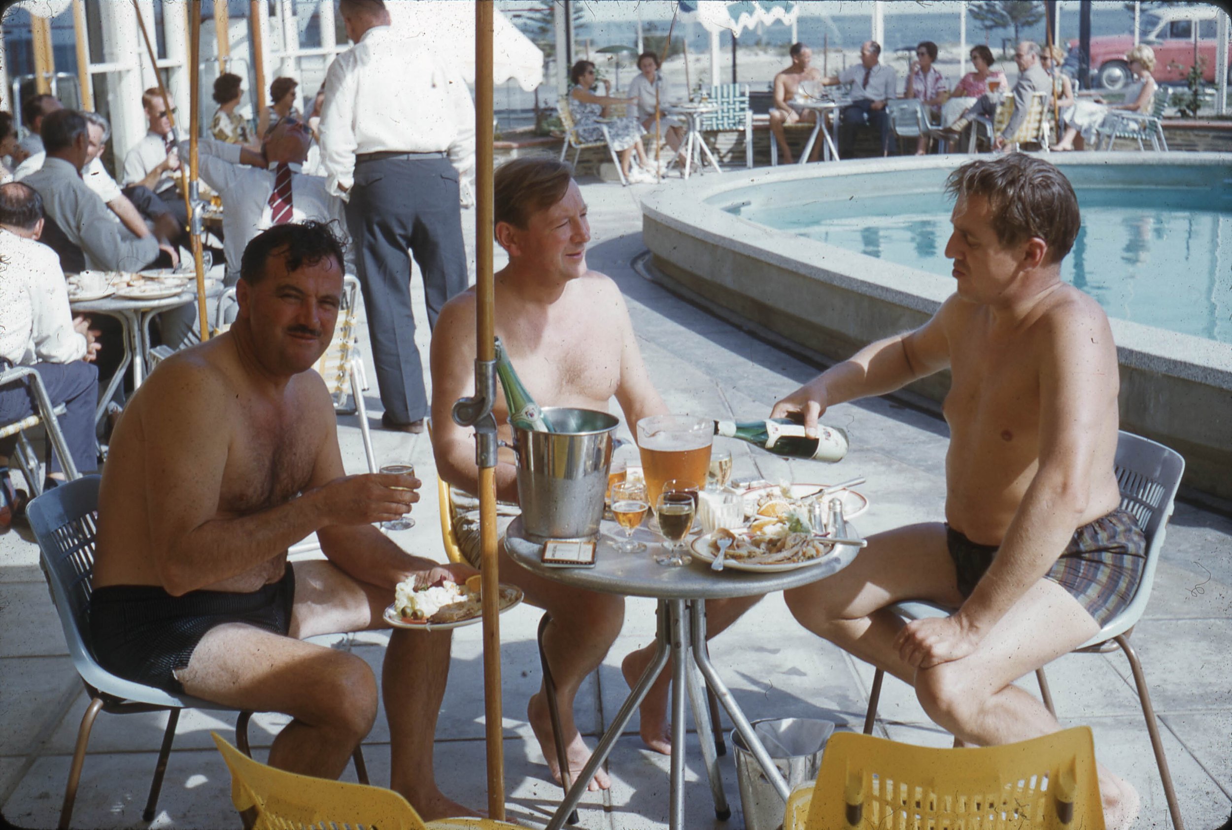 Three men sitting at a table outdoors, enjoying drinks and food, with a pool and cityscape in the background.