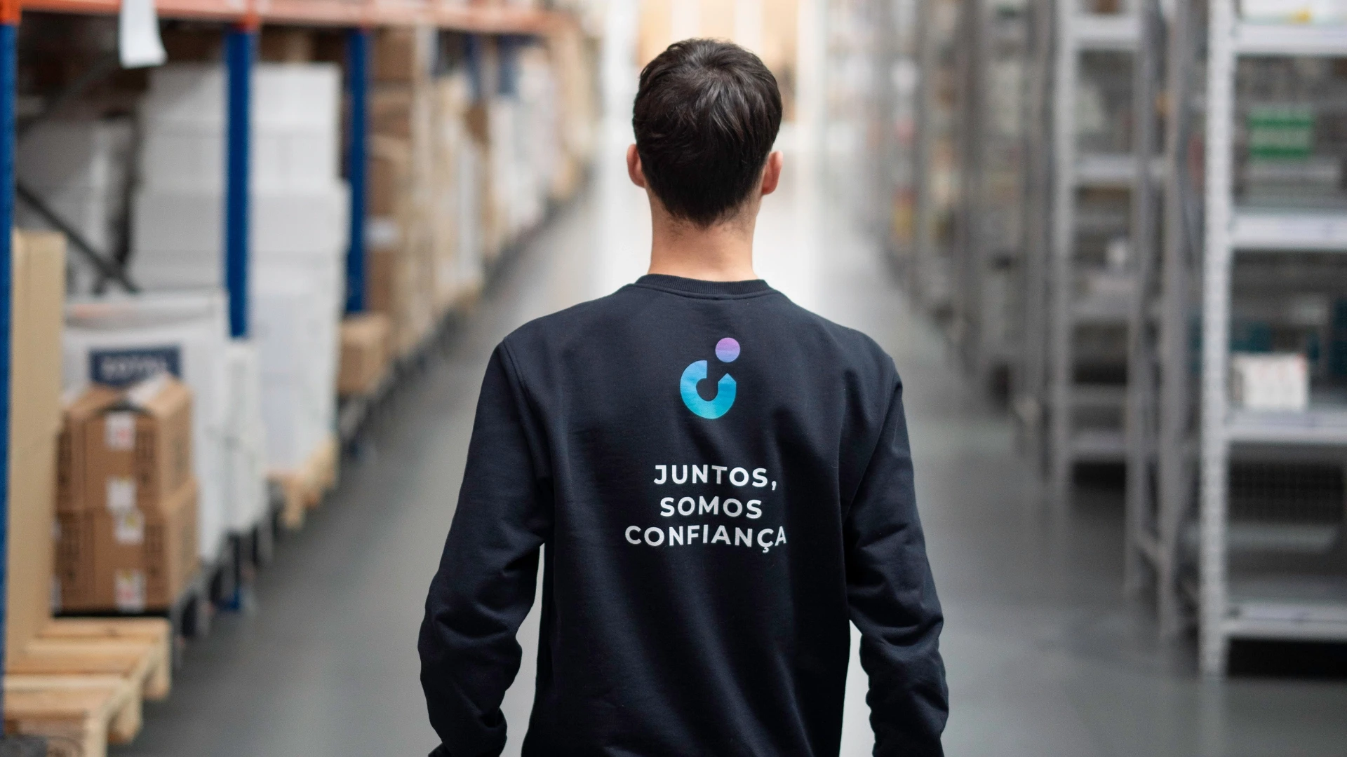 Pessoa de costas em um armazém de distribuição, vestindo uma camiseta com a frase 'Juntos, somos confiança', promovendo segurança e colaboração logística.