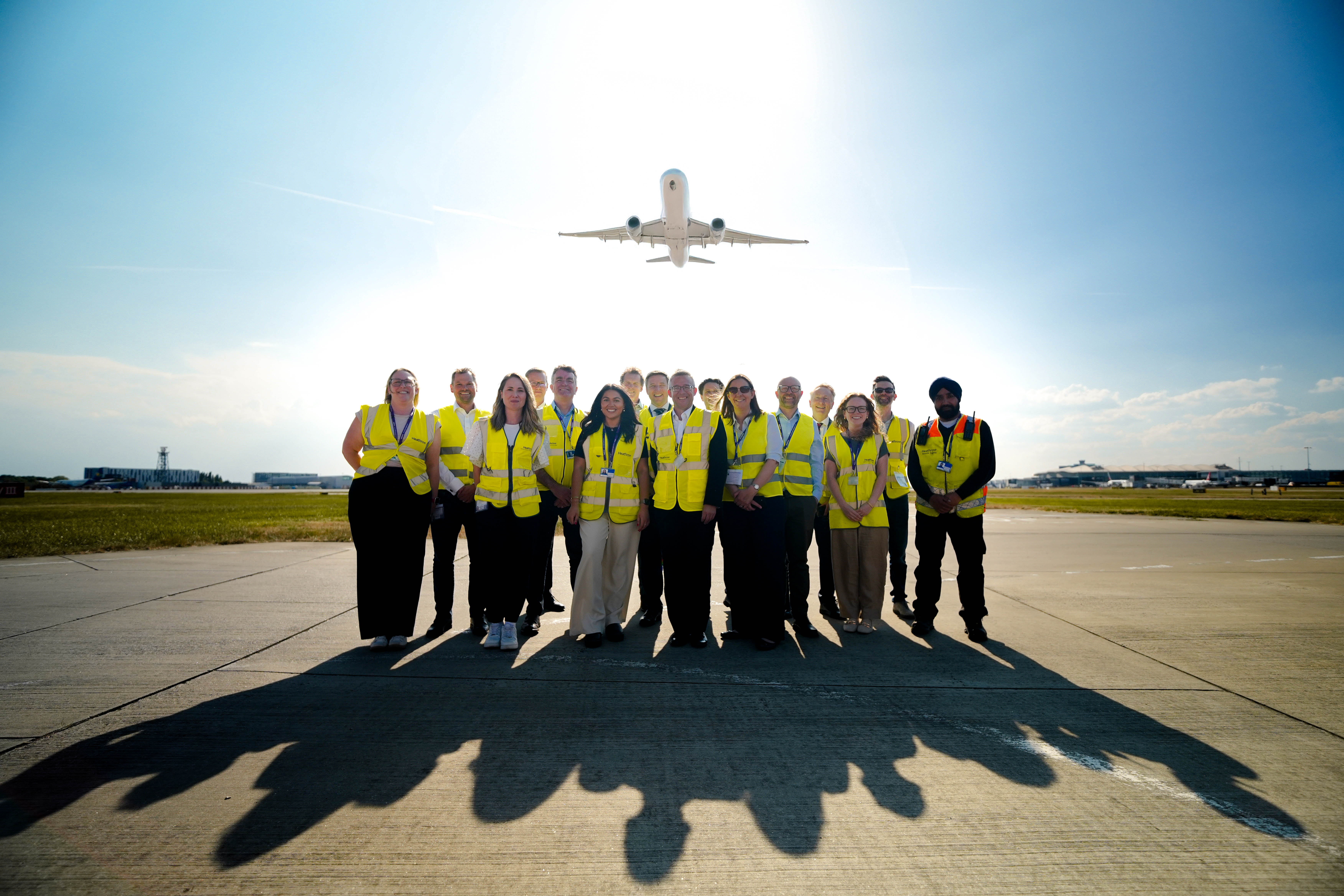 A group of approximately 15 people, many wearing yellow high-visibility vests, stand together for a group photo on a sunny airport tarmac. Directly centered above them in the bright blue sky, an airplane is in mid-flight, creating a dramatic backdrop.