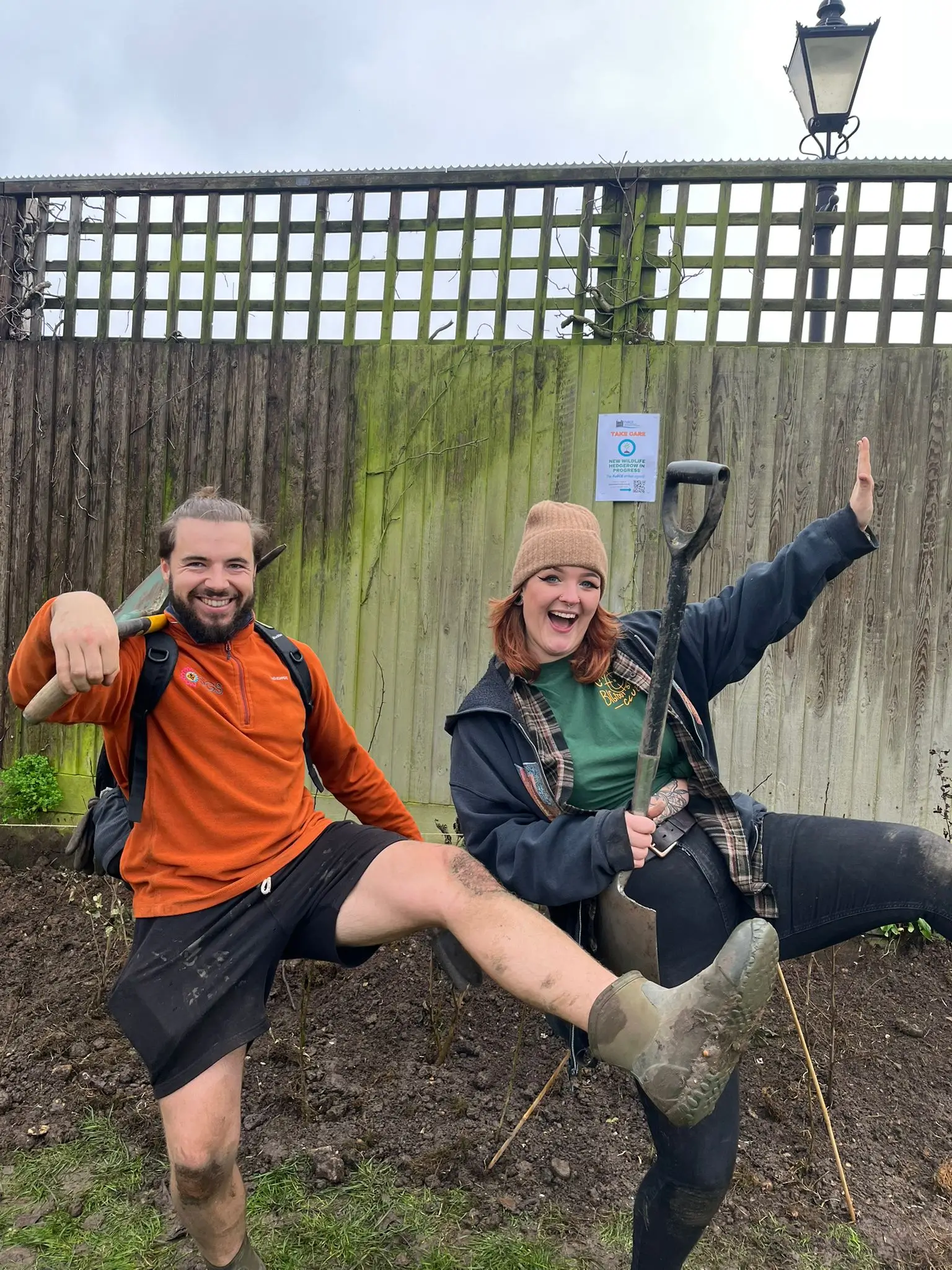 Two people pose playfully against a wooden fence, one wearing an orange shirt and the other in a black hoodie.