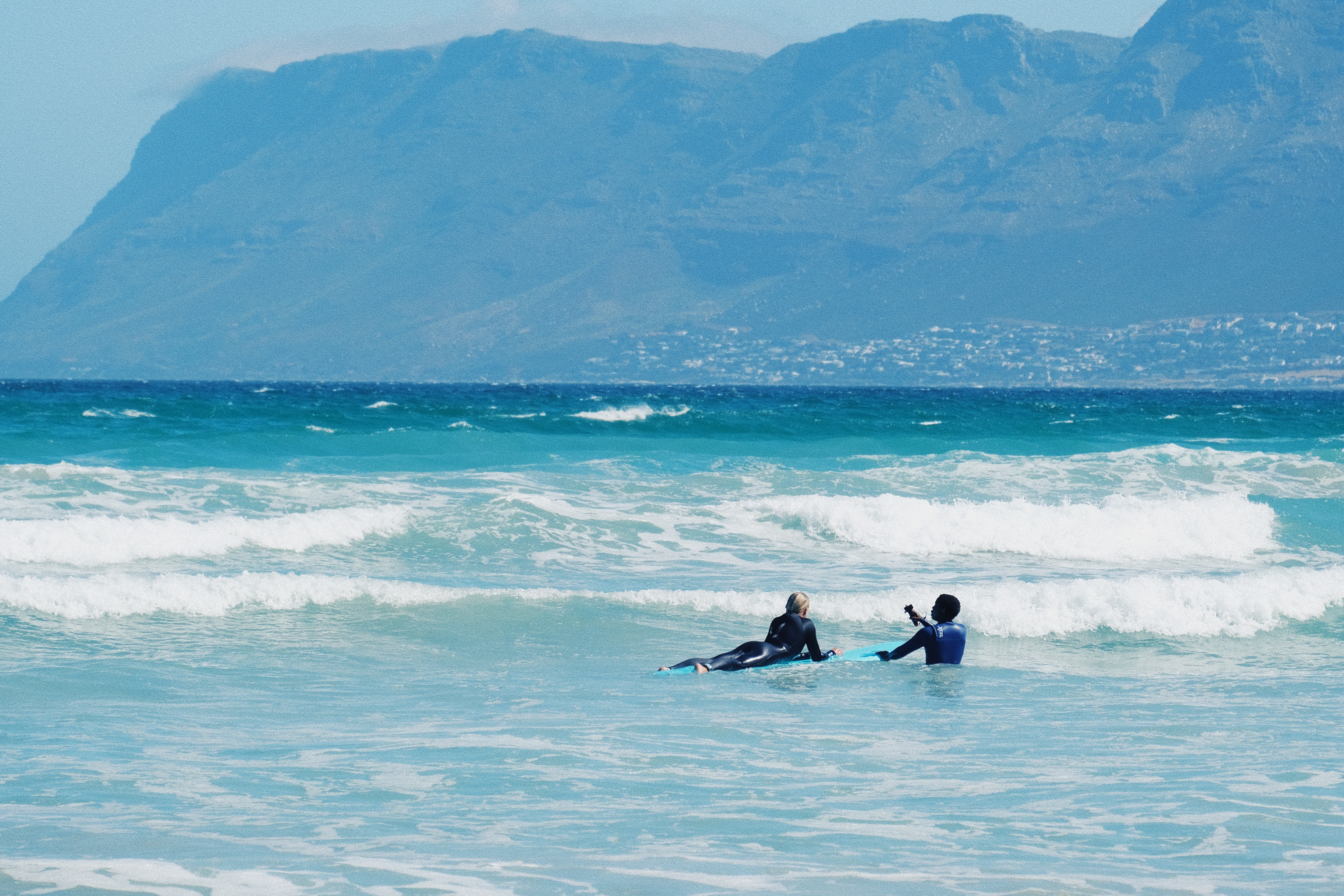 A surf student is laying on her surfboard while her surf coach explains her something, in the background you see beautiful Cape Town