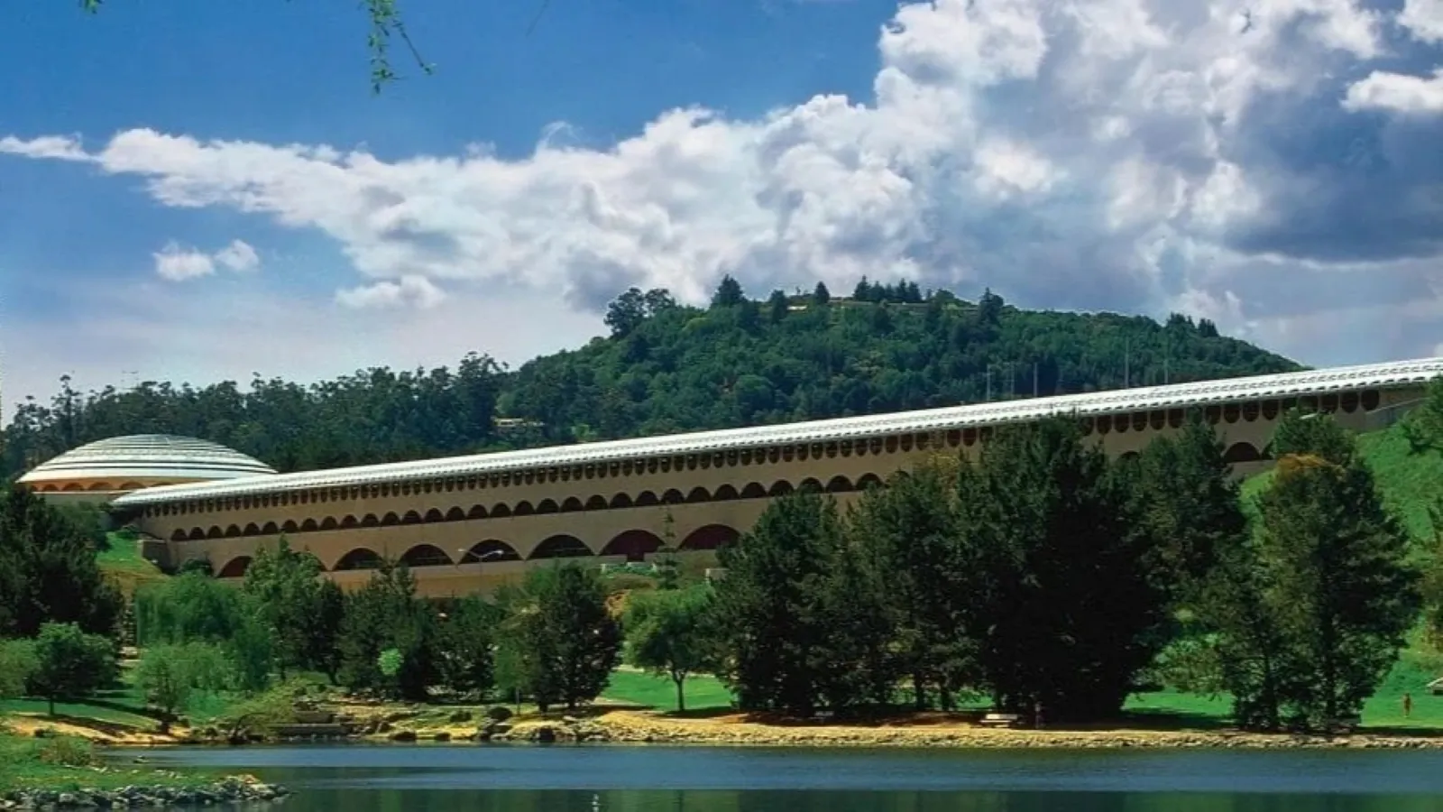 A large, modern architectural structure with a series of repeating arches stands amidst lush greenery and a serene lake, with a wooded hill and partly cloudy sky in the background, showcasing a harmonious blend of nature and design.