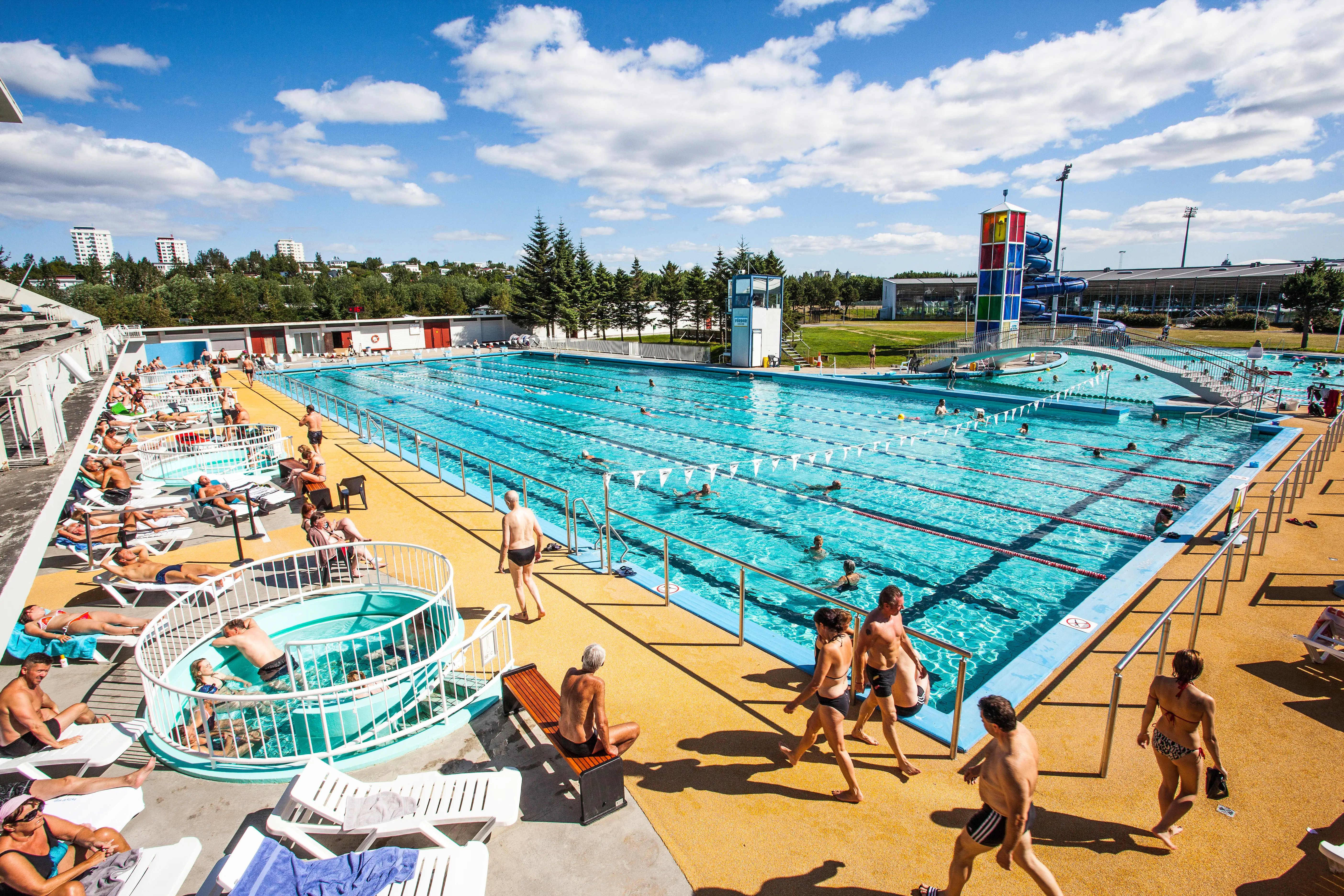Swimmers and sunbathers enjoying the large outdoor geothermal pool at Laugardalslaug in Reykjavík on a bright summer day, with lap lanes, hot tubs, and families relaxing around the pool deck.