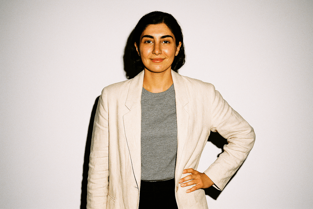 Confident individual in a beige blazer and gray shirt, standing against a plain white background with a poised expression.
