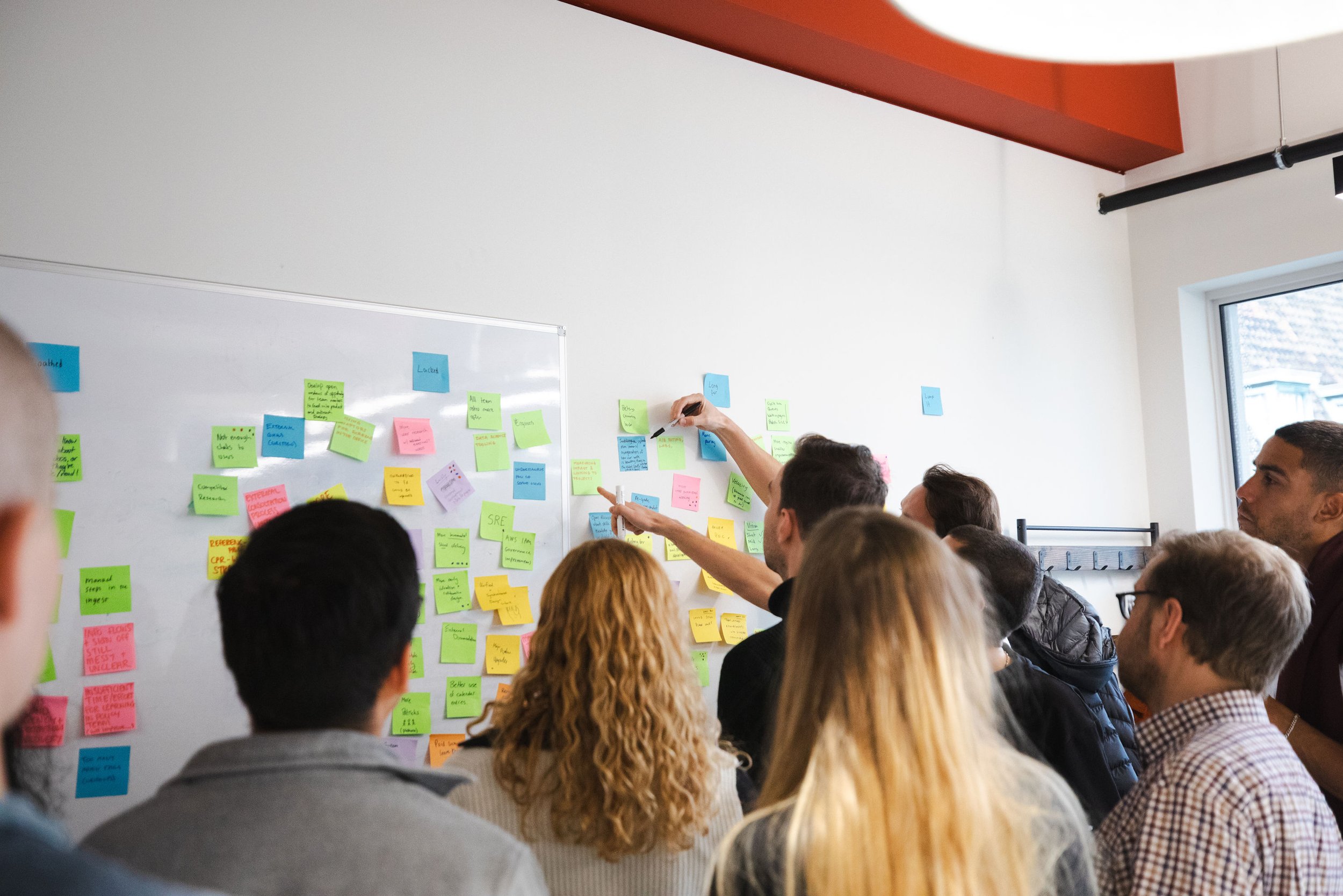 A team gathered around a whiteboard covered in colourful sticky notes during a workshop session, with one person reaching up to add or point to notes on the wall beyond the board.