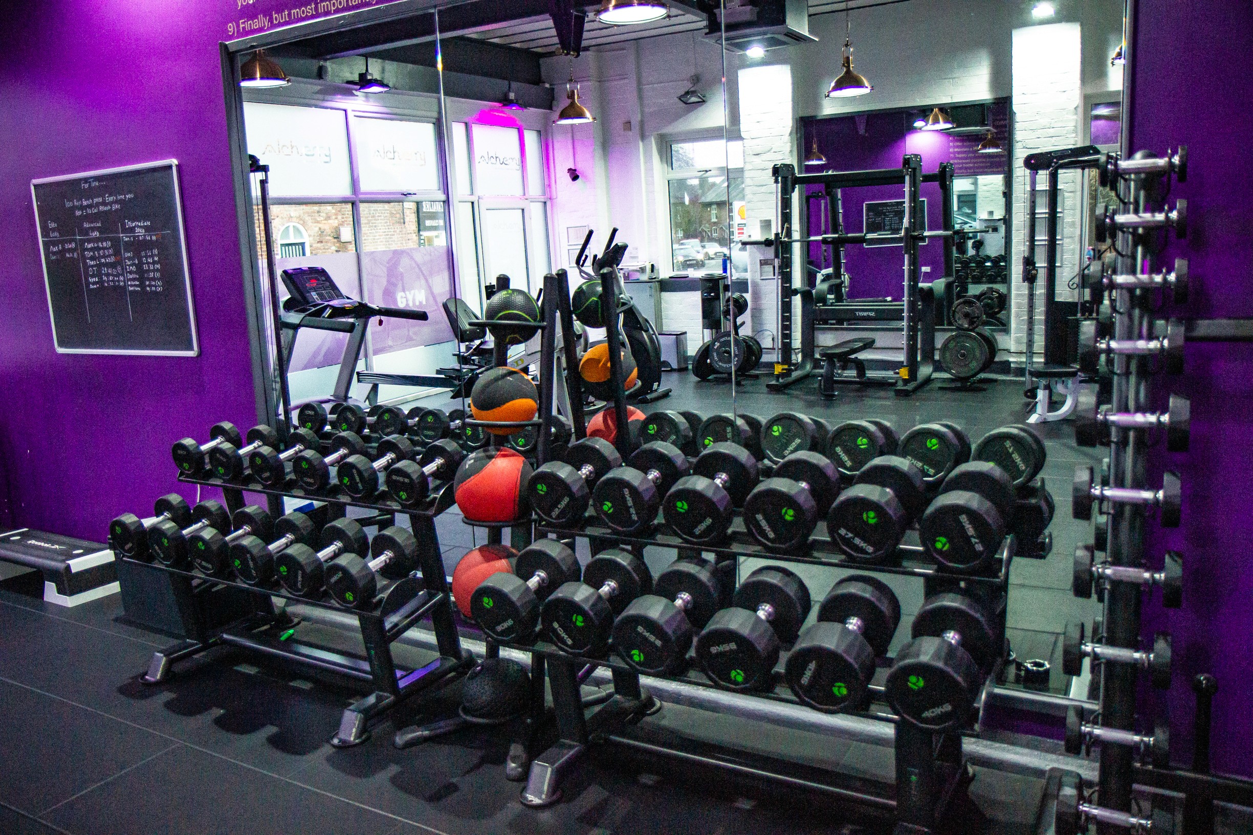 Alchemy PT gym interior with rows of dumbbells, weight machines, benches, purple accent walls, and a large mirror.