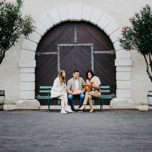 三个人坐在长椅上，长椅位于石墙中的拱形木门前，两边各有一棵树。