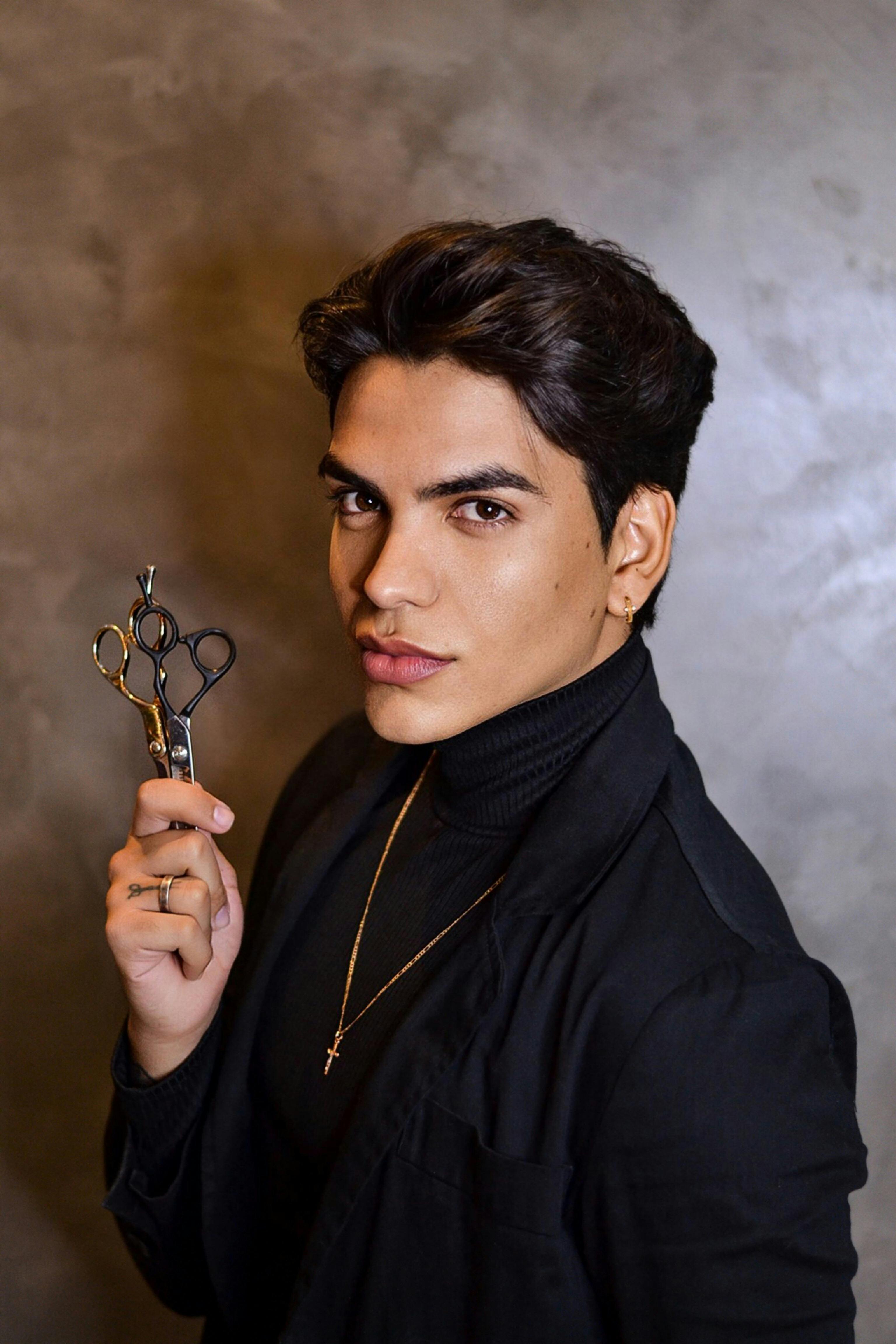 A young man with dark hair, a black jacket, and a gold necklace holds hair scissors, looking at the camera.