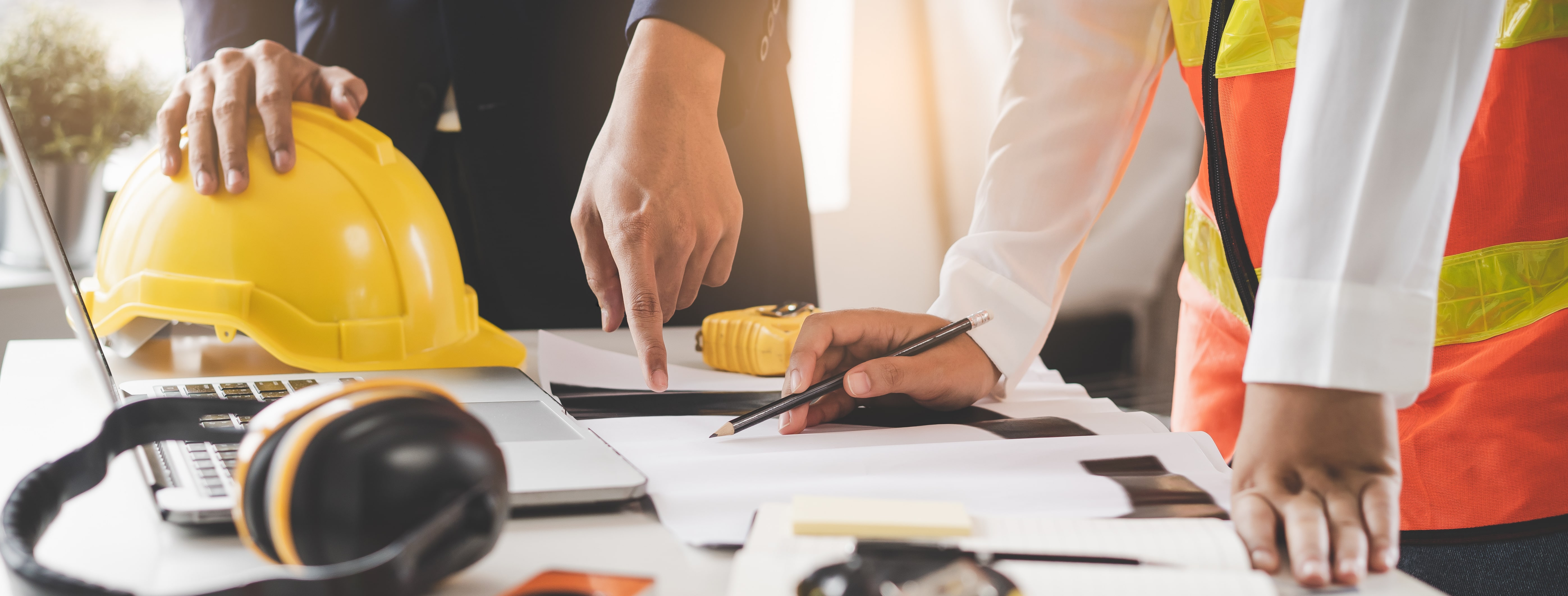Two professionals, one in a suit and the other in a safety vest, reviewing construction plans on a desk with a yellow hard hat, laptop, and safety gear.