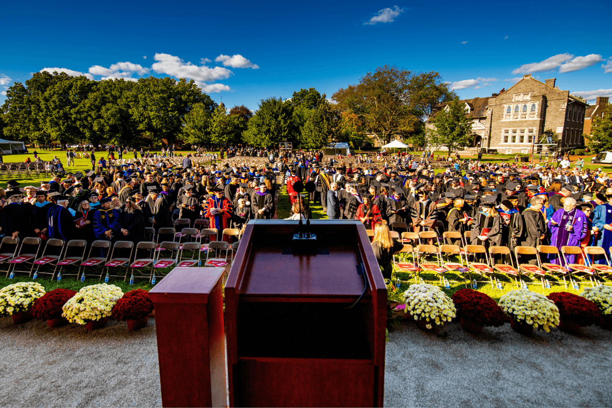 View from the podium looking out on a commencement crowd with blue sky, trees, and academic building in the background