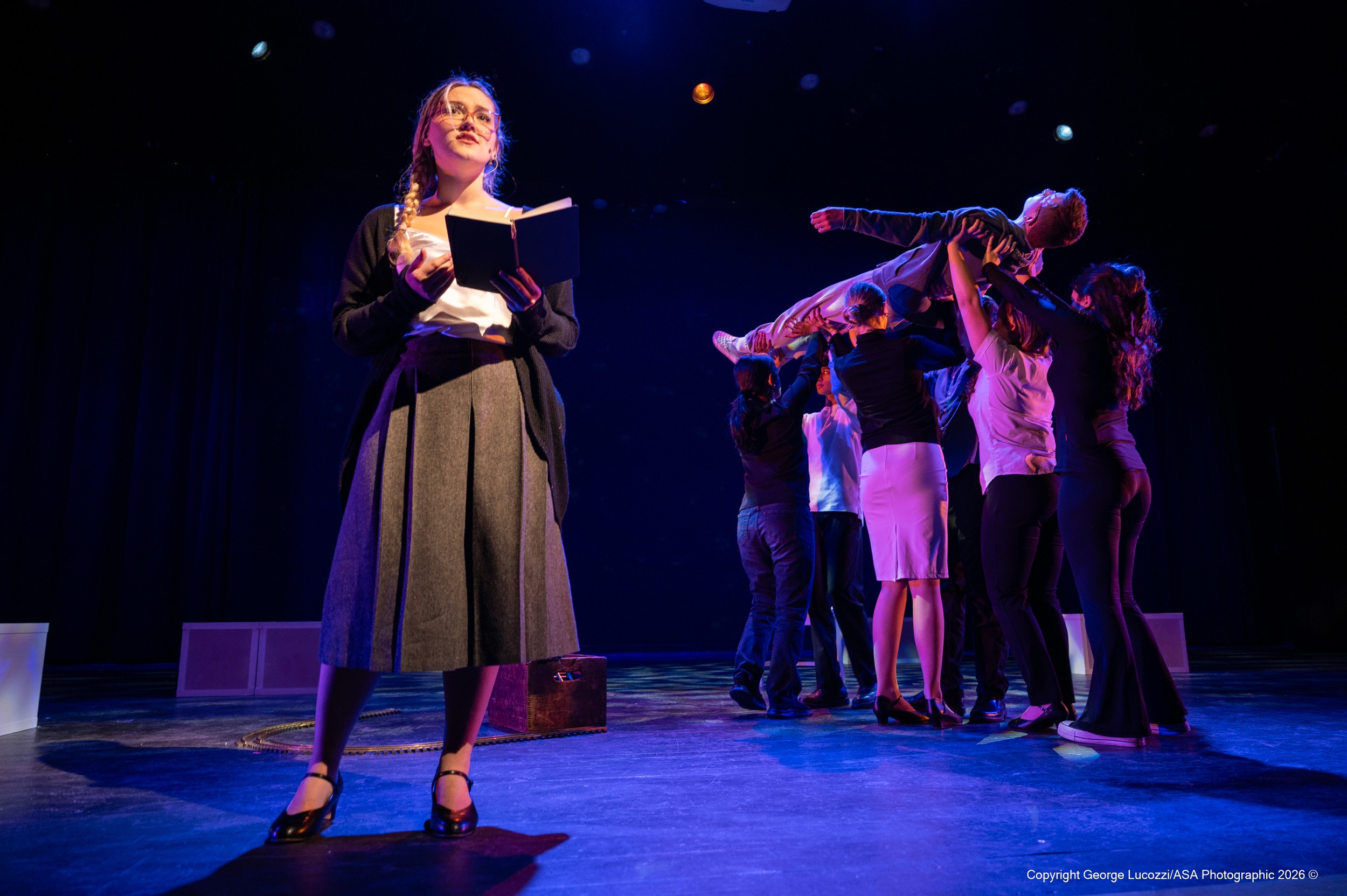 Siobhan stands downstage in a focused followspot reading from a book, while upstage the “Voices” lift Christopher horizontally; dramatic blue sidelight and warm top light sculpt the ensemble against a field of subtle star-like gobo dots.