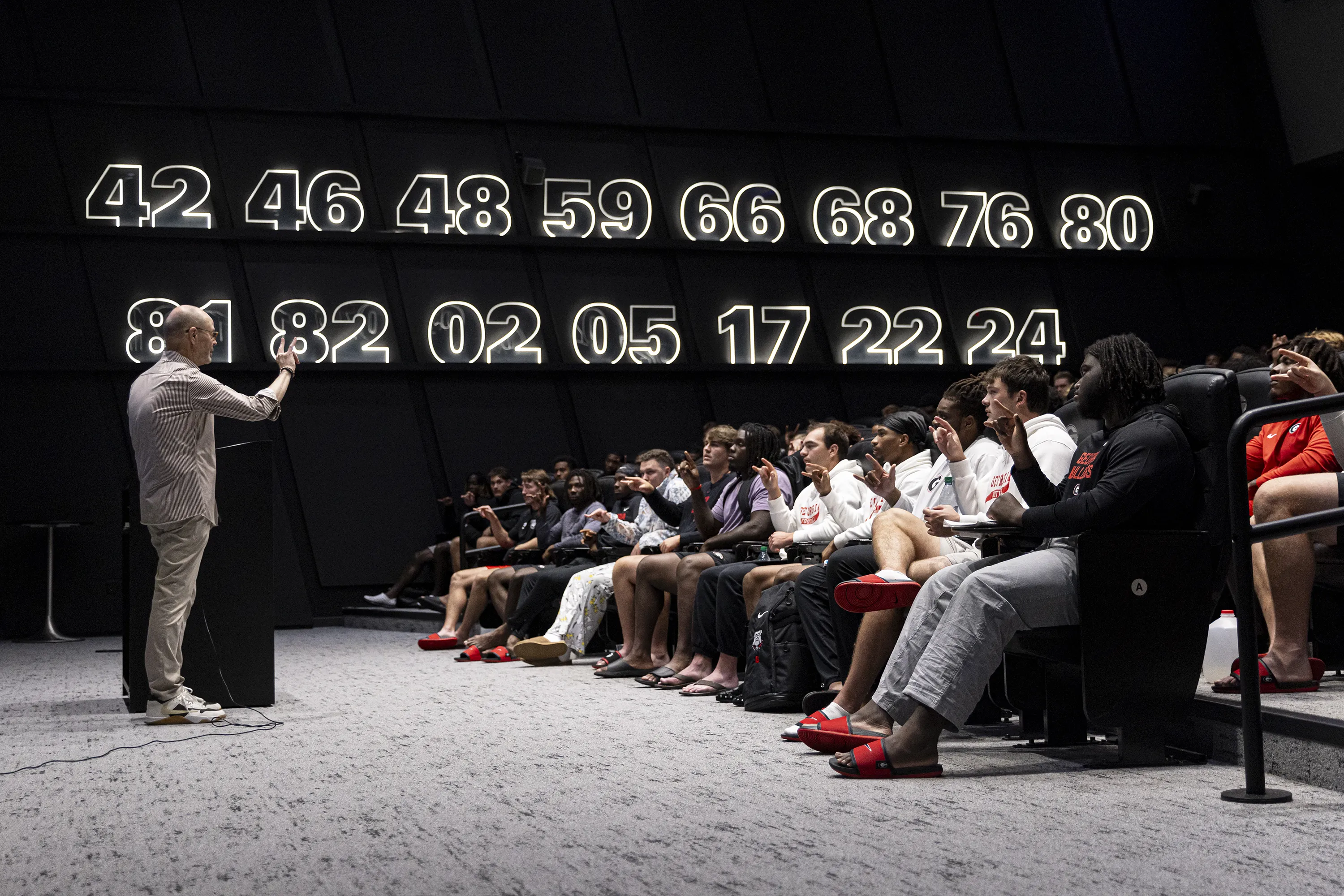 A person stands at a podium speaking to a group of seated people in a dark room with large, illuminated numbers on the back wall.