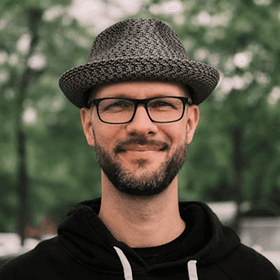 A man wearing a gray fedora and black-rimmed glasses smiles outdoors in front of blurred green trees, dressed in a black hoodie.