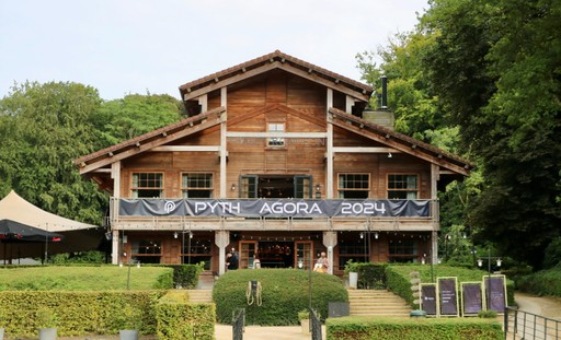A large wooden lodge-style building surrounded by trees, displaying a black banner that reads “PYTH AGORA 2024” across the upper balcony. The entrance area has trimmed hedges, steps leading up, and several vertical event boards on the right side.