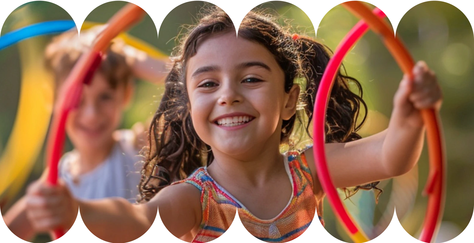 Uma menina sorrindo com cabelo escuro preso, brincando e segurando arcos coloridos em um ambiente externo ensolarado. Outra criança está desfocada ao fundo.